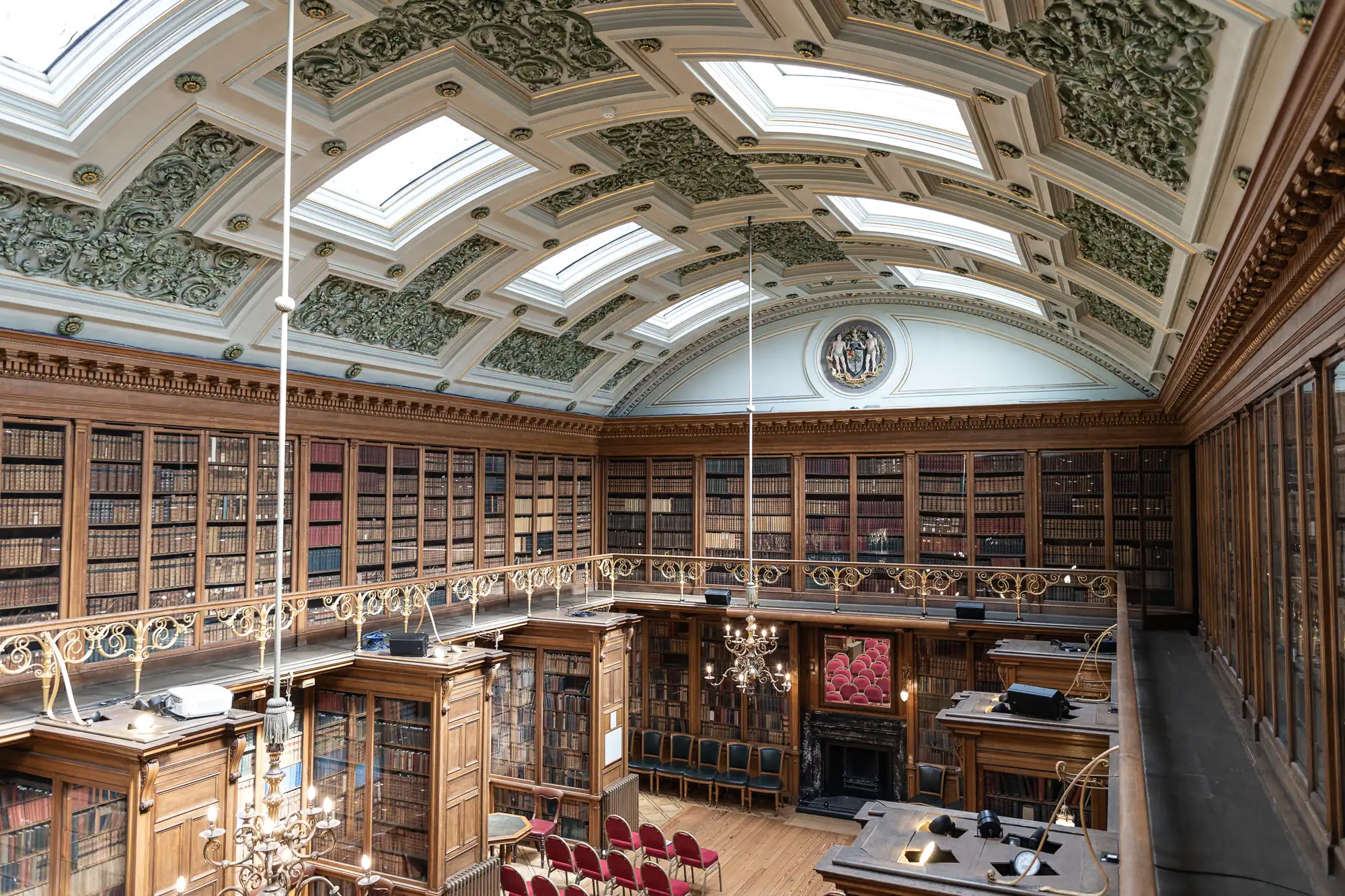 Ornate library interior with wooden bookshelves filled with books, a decorative ceiling with skylights, and a section with red chairs facing a lectern and fireplace.