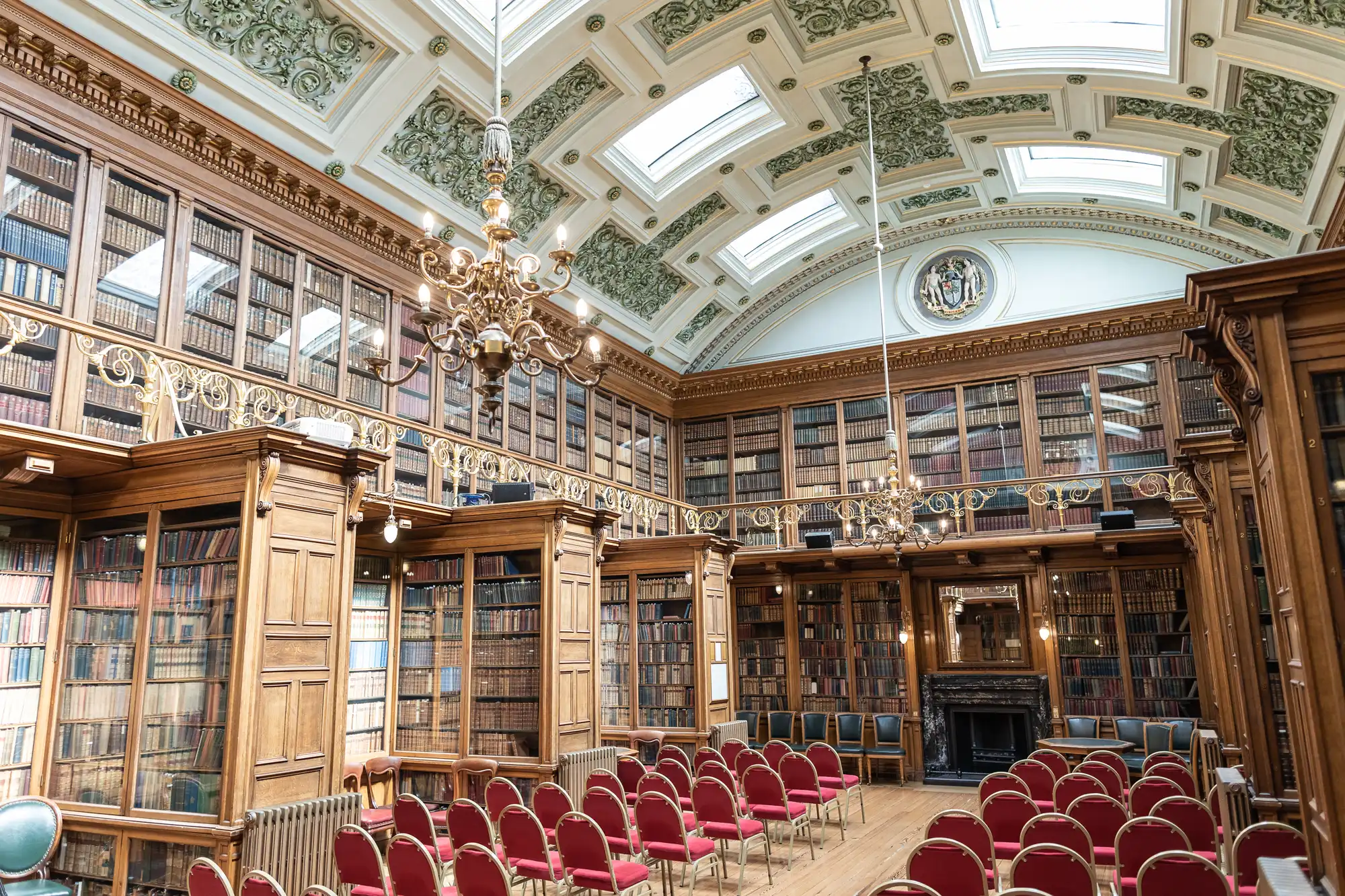 A grand library room with ornate ceiling, wooden bookshelves filled with books, and red chairs arranged in rows facing a fireplace and podium. Chandeliers hang from the high ceiling.