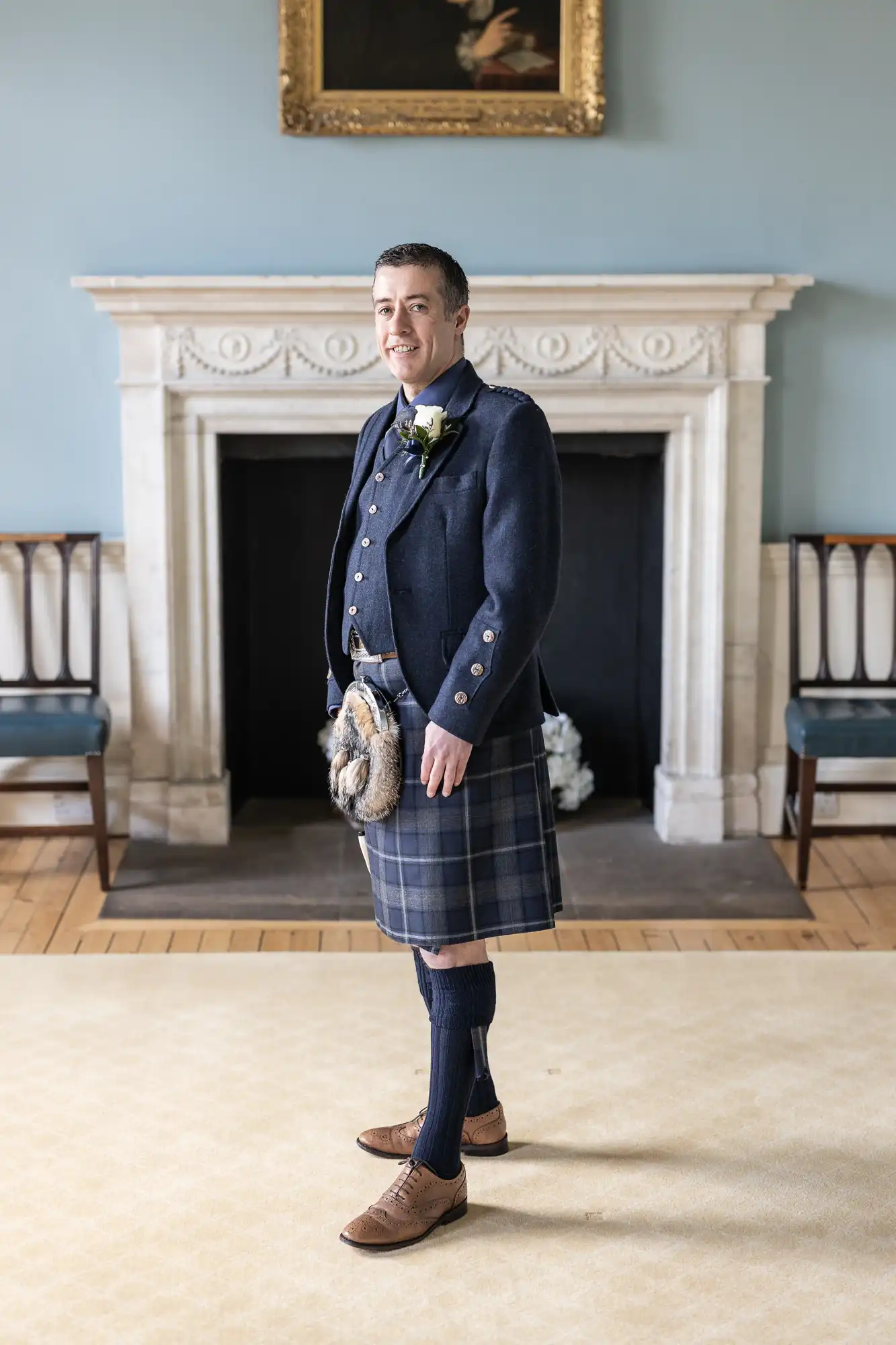 A man stands indoors wearing a traditional Scottish kilt outfit in front of a white fireplace, posing for the camera.