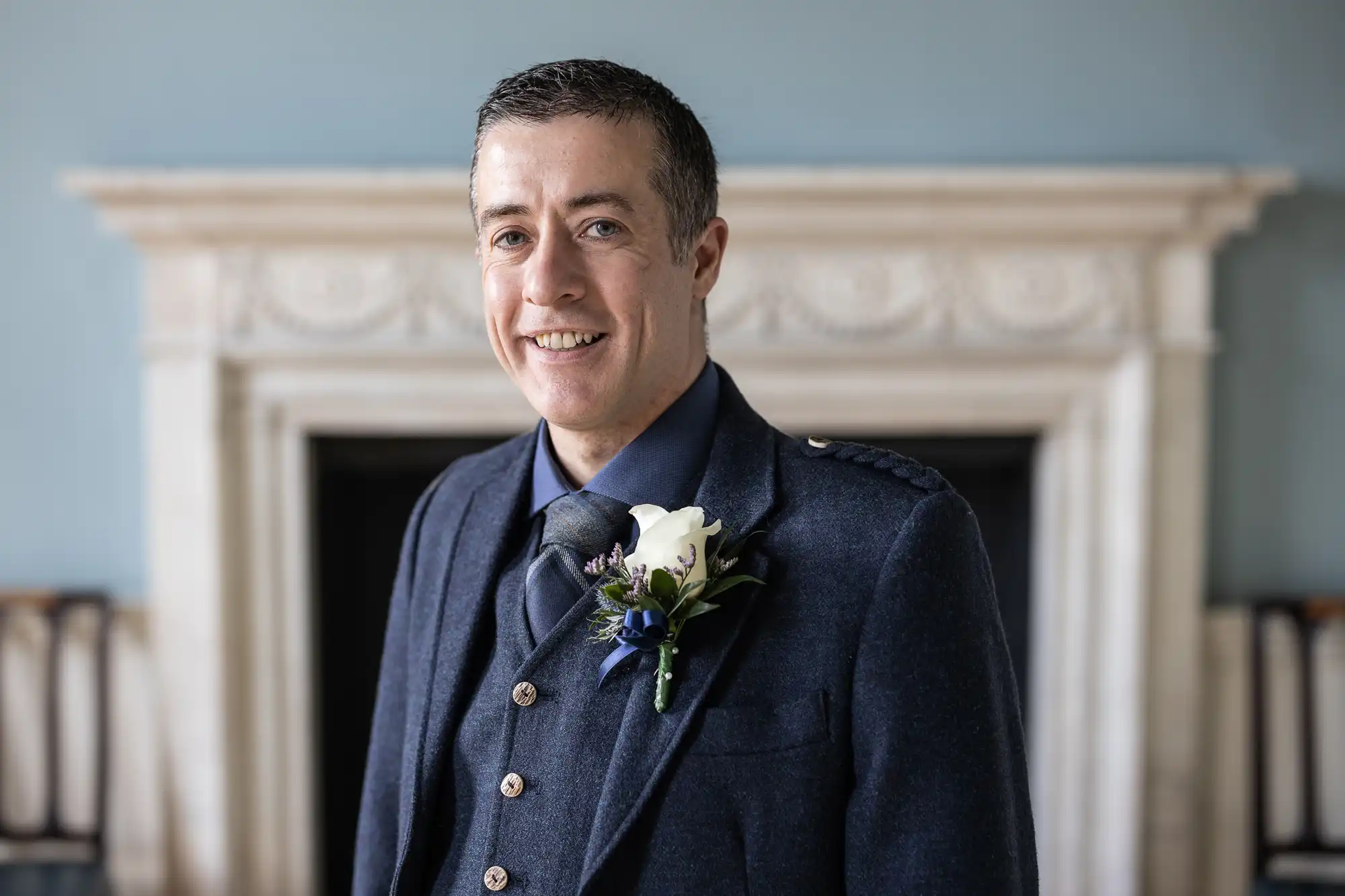 A man in a blue suit with a flower boutonniere stands in front of an ornate fireplace, smiling at the camera.