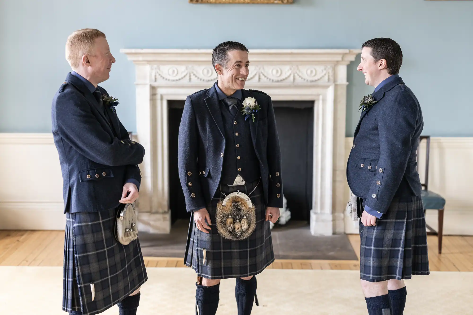 Three men, dressed in traditional Scottish kilts and jackets, stand and laugh together in front of a fireplace in a well-lit room.