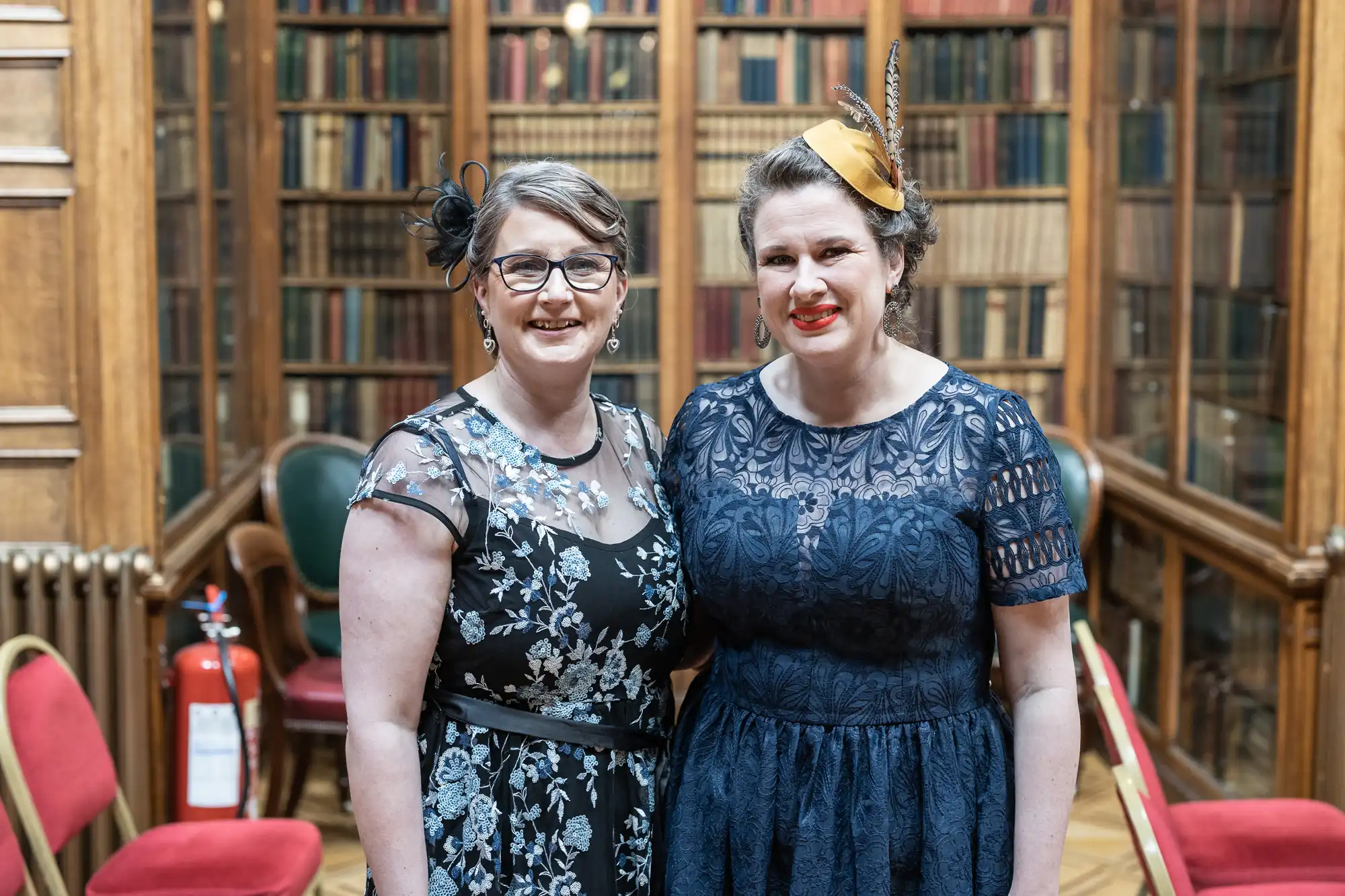 Two women, each in an evening dress, stand side by side in a room with bookshelves and chairs. One woman wears glasses and a flowered dress. The other woman wears a fascinator and a lace dress.