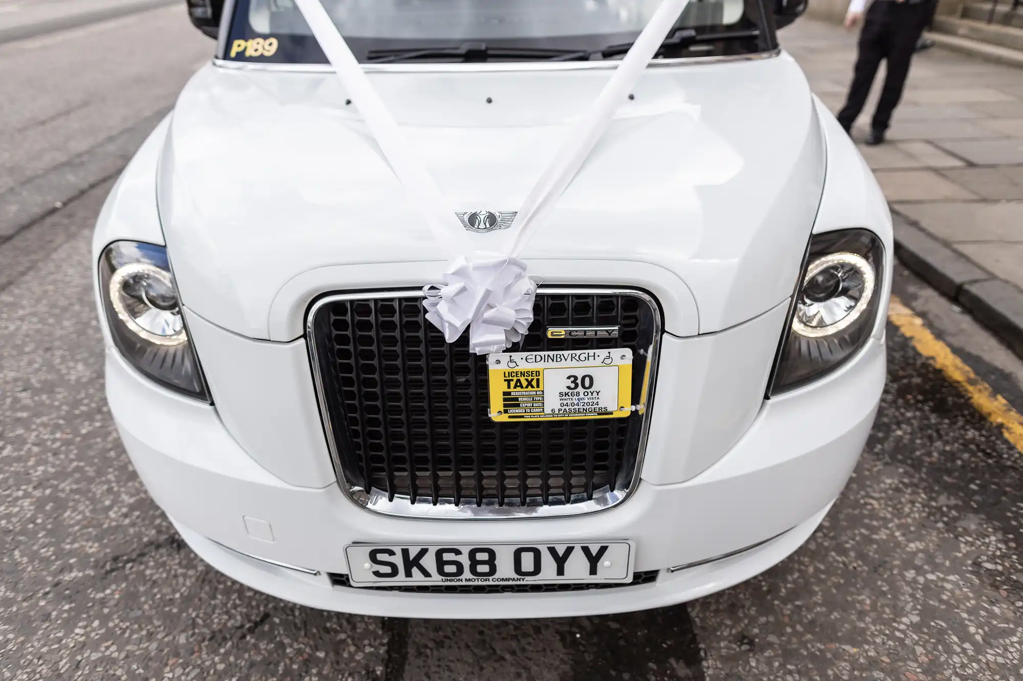 A white taxi decorated with a white ribbon on the front grille, displaying a license plate SK68 OYY and a taxi permit for Edinburgh, parked on a street.