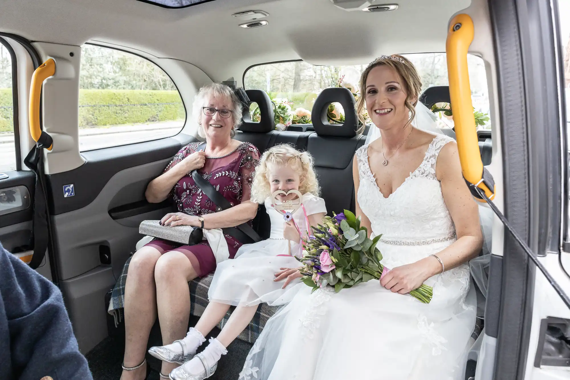 A bride, a young girl, and an elderly woman sit inside a vehicle. The bride holds a bouquet, the young girl wears a white dress and holds a heart-shaped wand, while the elderly woman smiles.
