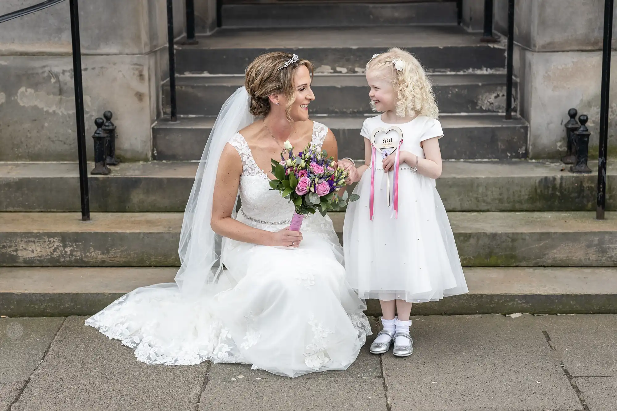 A bride in a white wedding dress sits on stone steps holding a bouquet while smiling at a young girl in a white dress holding a "2017" sign.