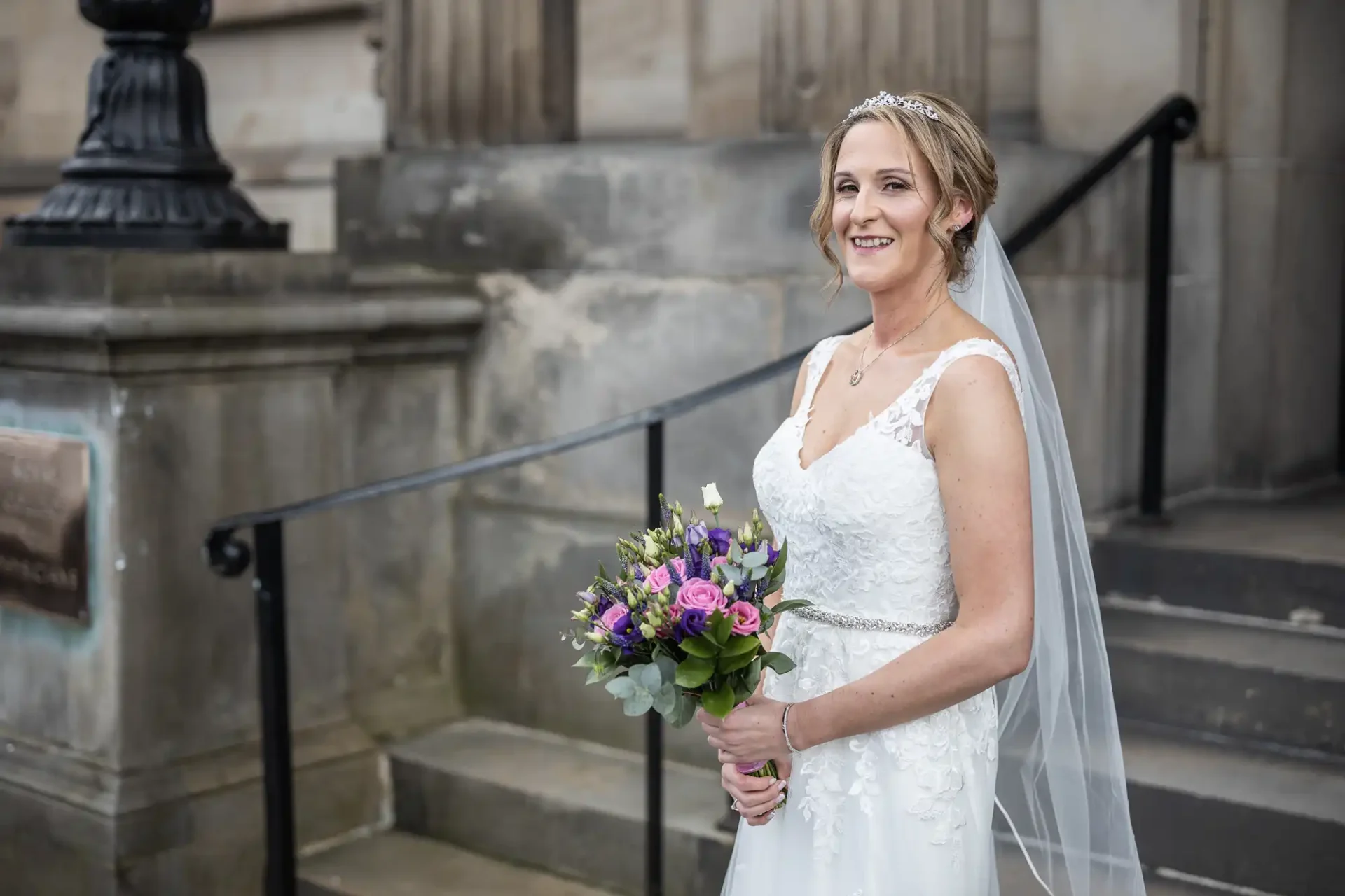 A bride in a white wedding dress holds a bouquet of flowers and smiles while standing outside a stone building with steps.