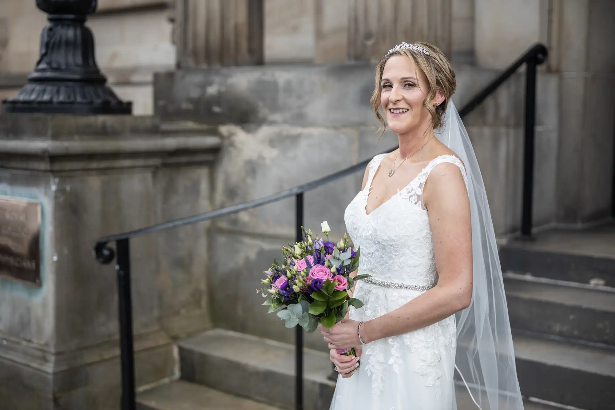 A bride in a white wedding dress holds a bouquet of flowers and smiles while standing outside a stone building with steps.