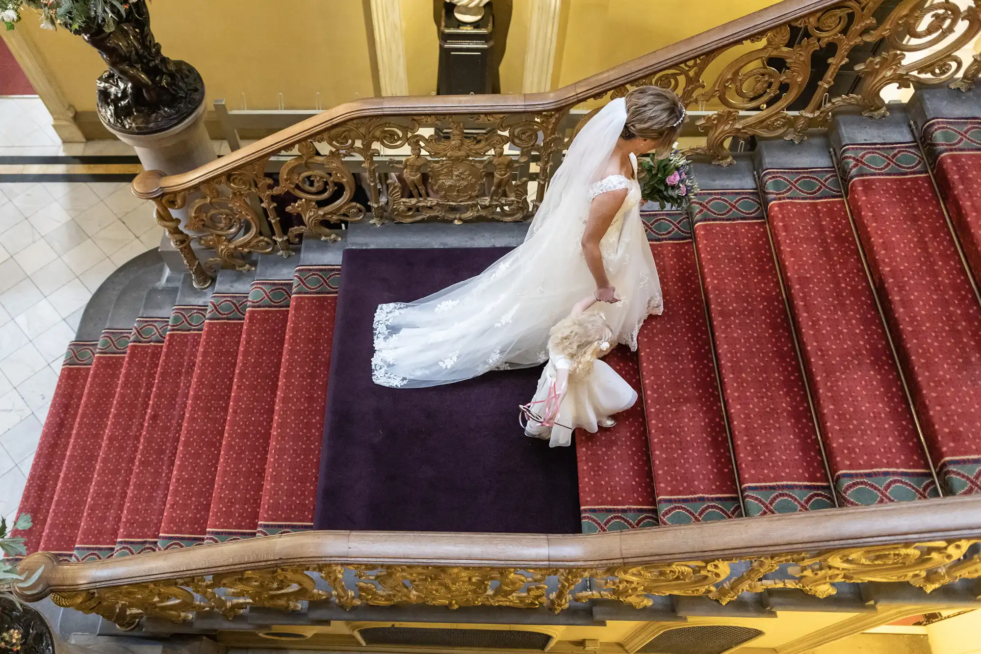 A bride in a white gown and veil walks up a grand staircase with a young flower girl, both holding hands. The staircase has red carpet and ornate golden railing.