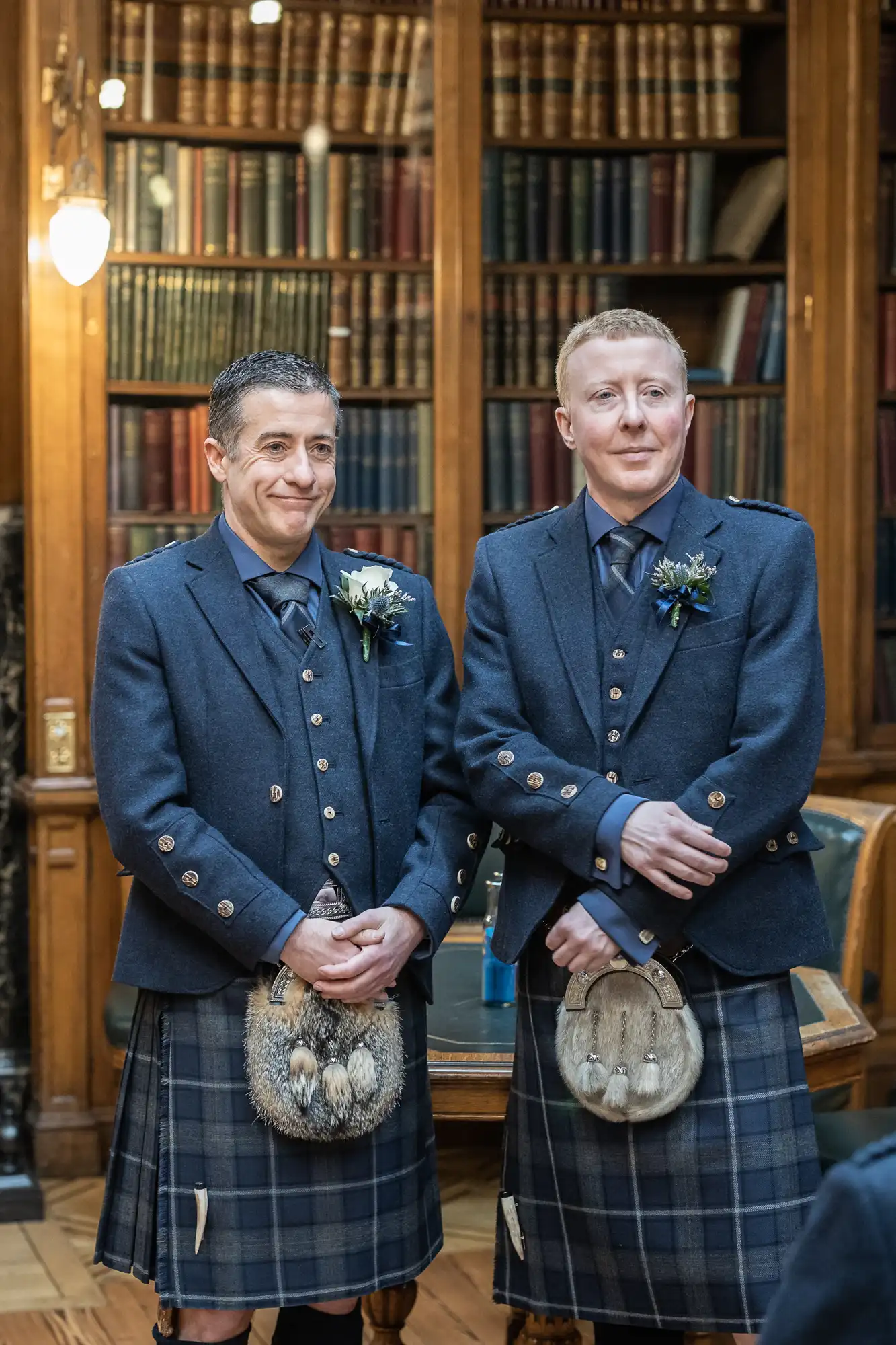 Two men in blue kilts stand side by side in a wood-paneled room with bookshelves in the background, both smiling.