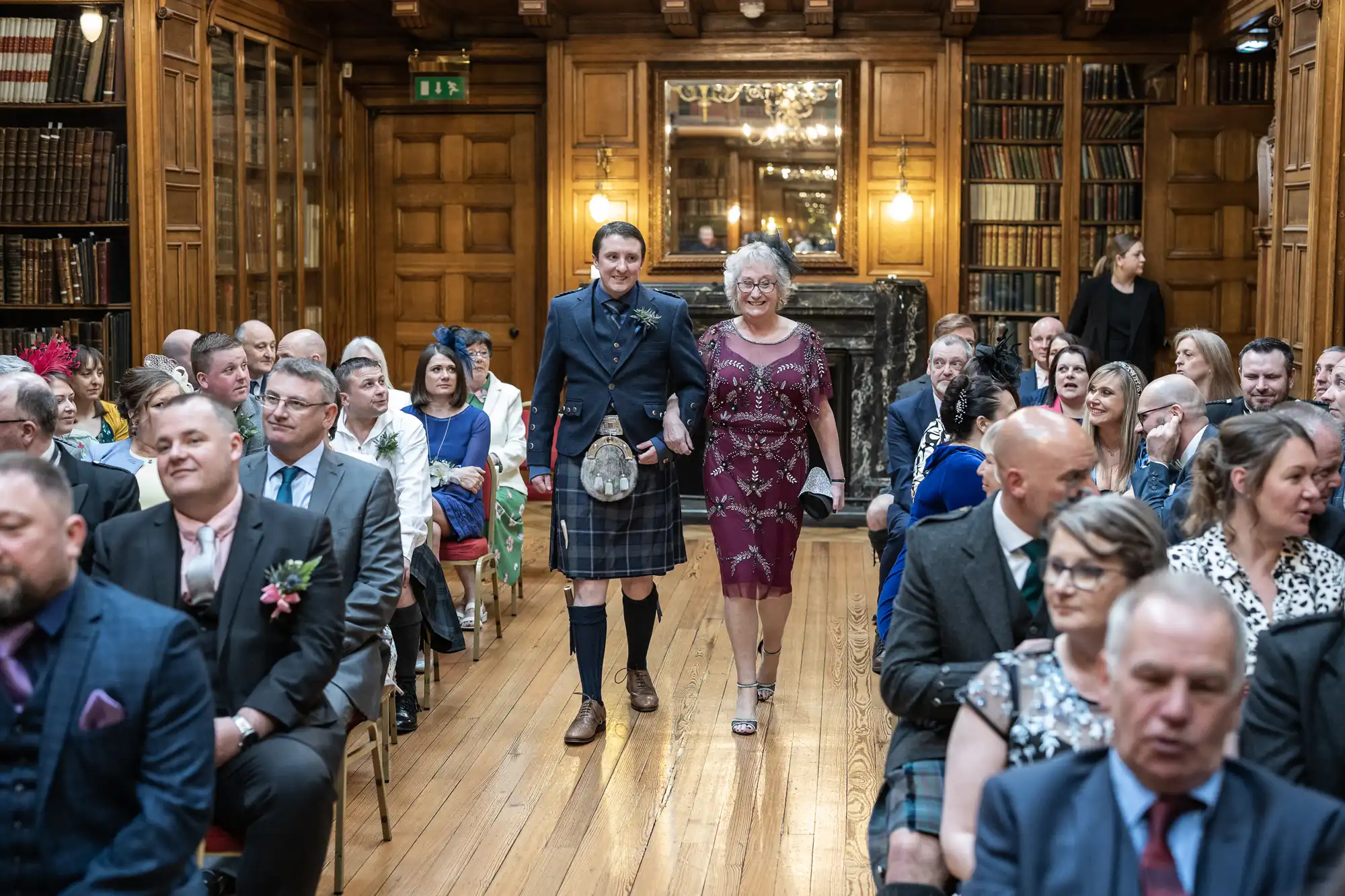 Two people walk down the aisle in a wood-paneled room filled with seated guests. The person on the left wears a kilt, and the person on the right wears a red dress.