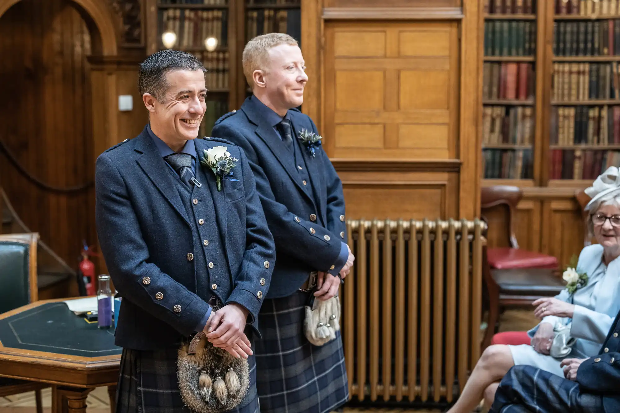 Two individuals wearing traditional Scottish kilts stand side by side, smiling in a wood-paneled room with bookshelves. An audience member is partially visible to the right.