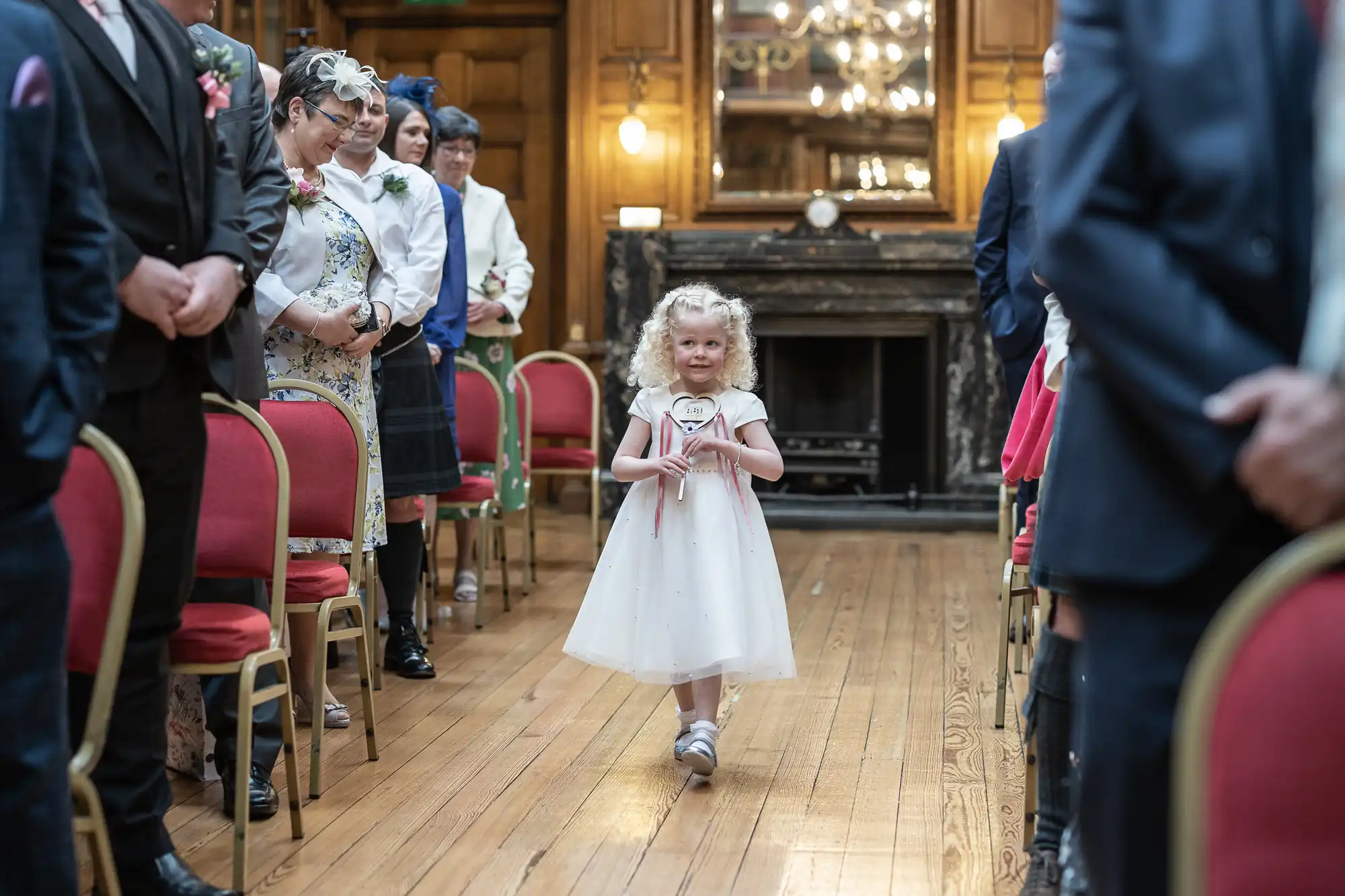 A young girl in a white dress walks down an aisle holding a sign at a wedding ceremony, with seated guests watching.