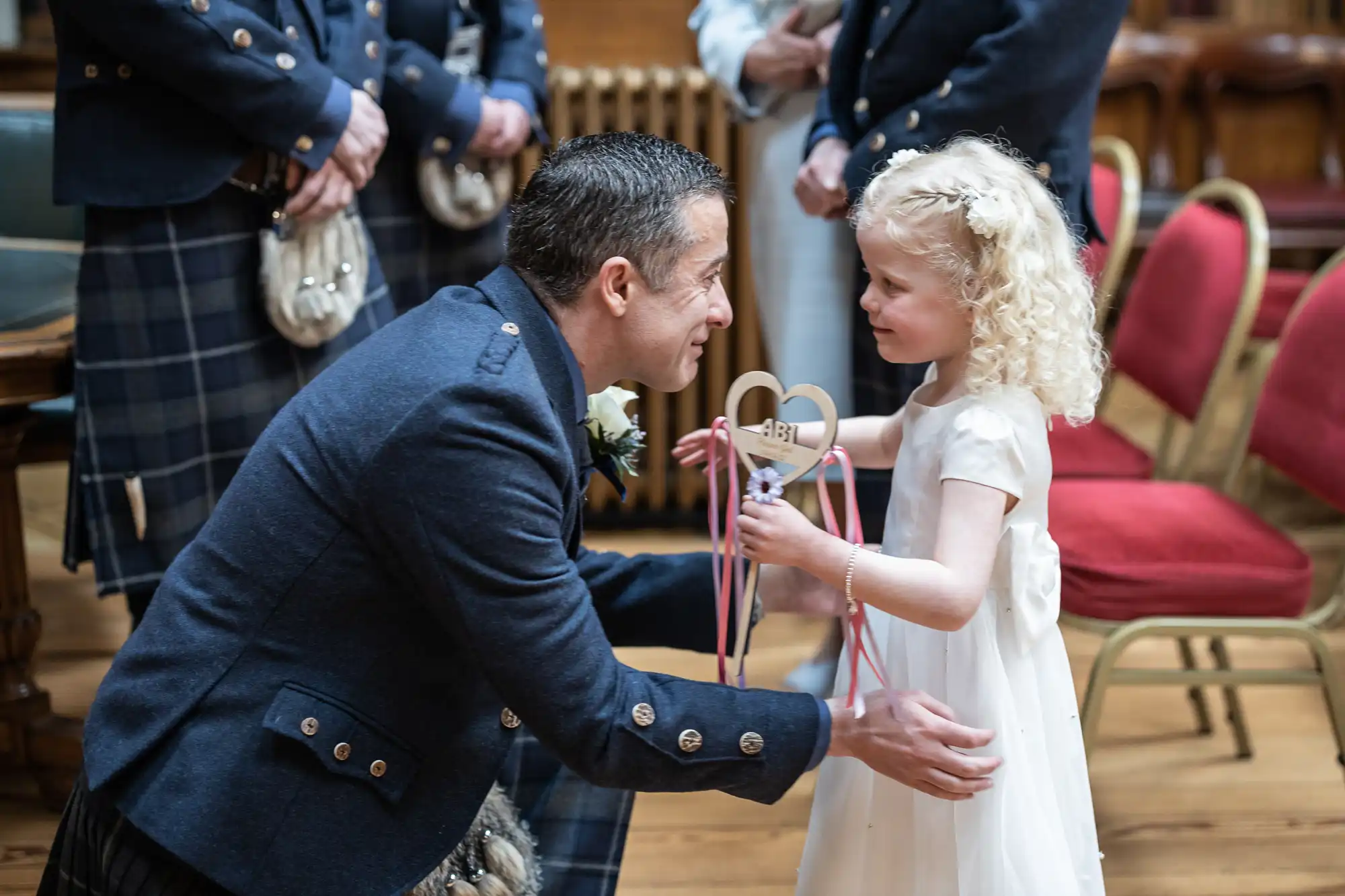 A man in a blue jacket kneels and smiles at a young girl in a white dress holding a wooden heart-shaped ring bearer box with ribbons. Both are about to present or receive something in a formal setting.