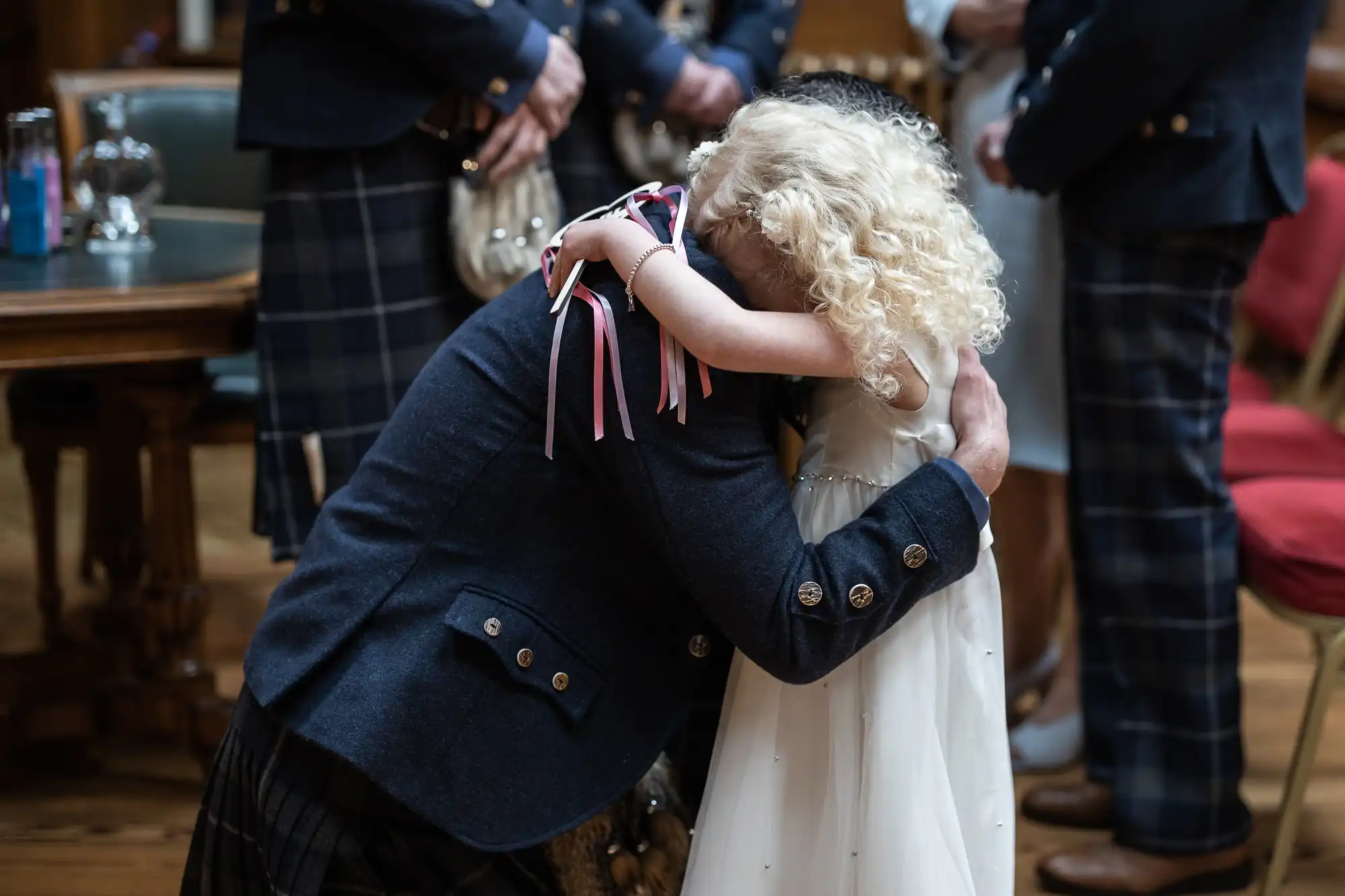 A man in traditional Scottish attire hugs a young girl with curly blonde hair during an event.