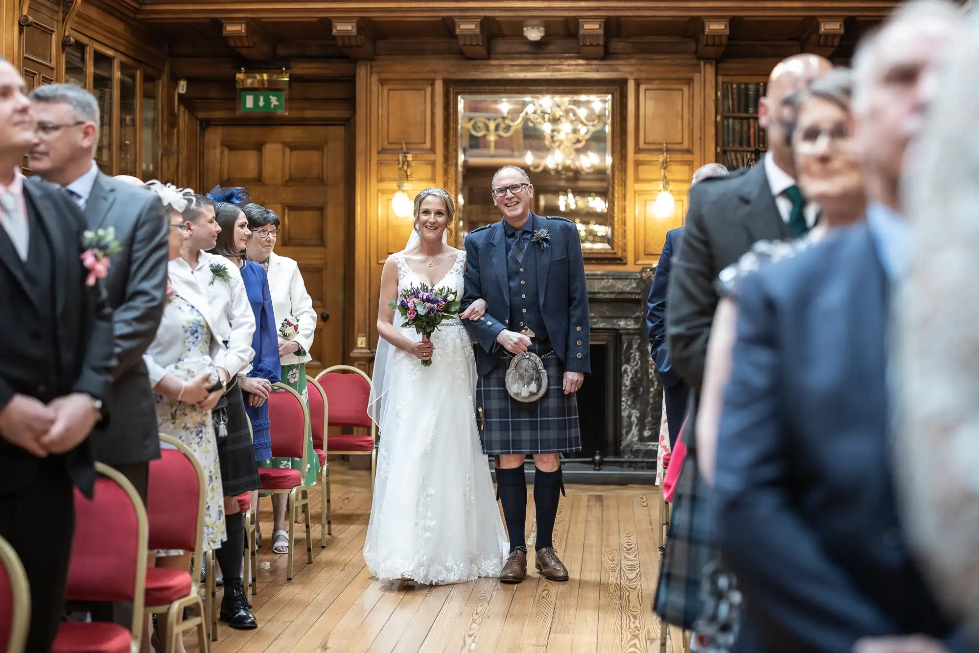 A bride walks down the aisle with a man in traditional Scottish attire, surrounded by seated guests in a wooden-paneled room.