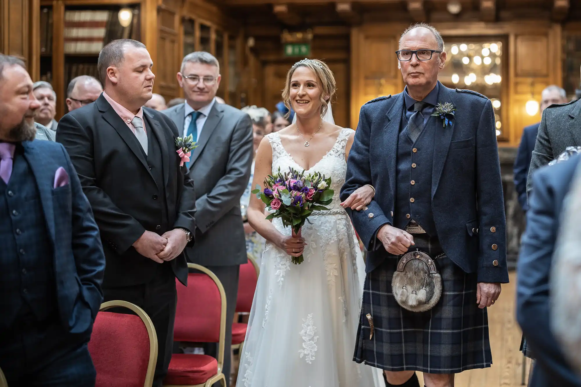 A bride holding a bouquet and smiling is walked down the aisle by an older man in a kilt, surrounded by standing men in suits inside a wooden-paneled room.