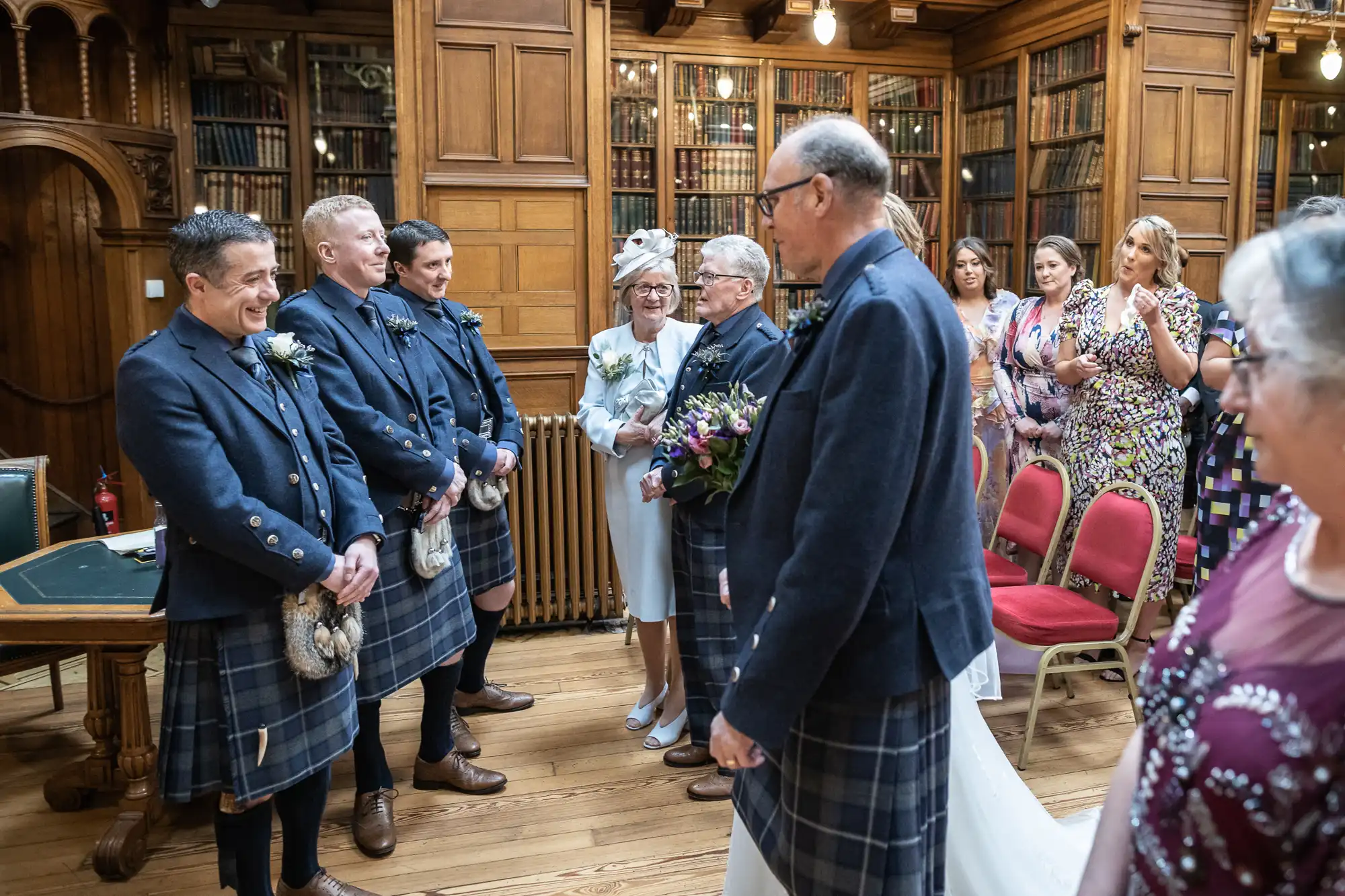 A wedding procession in a wood-paneled room: the groom, in a kilt, stands with groomsmen, waiting as the bride, carrying a bouquet, is escorted down the aisle by an elderly man. Seated guests look on.