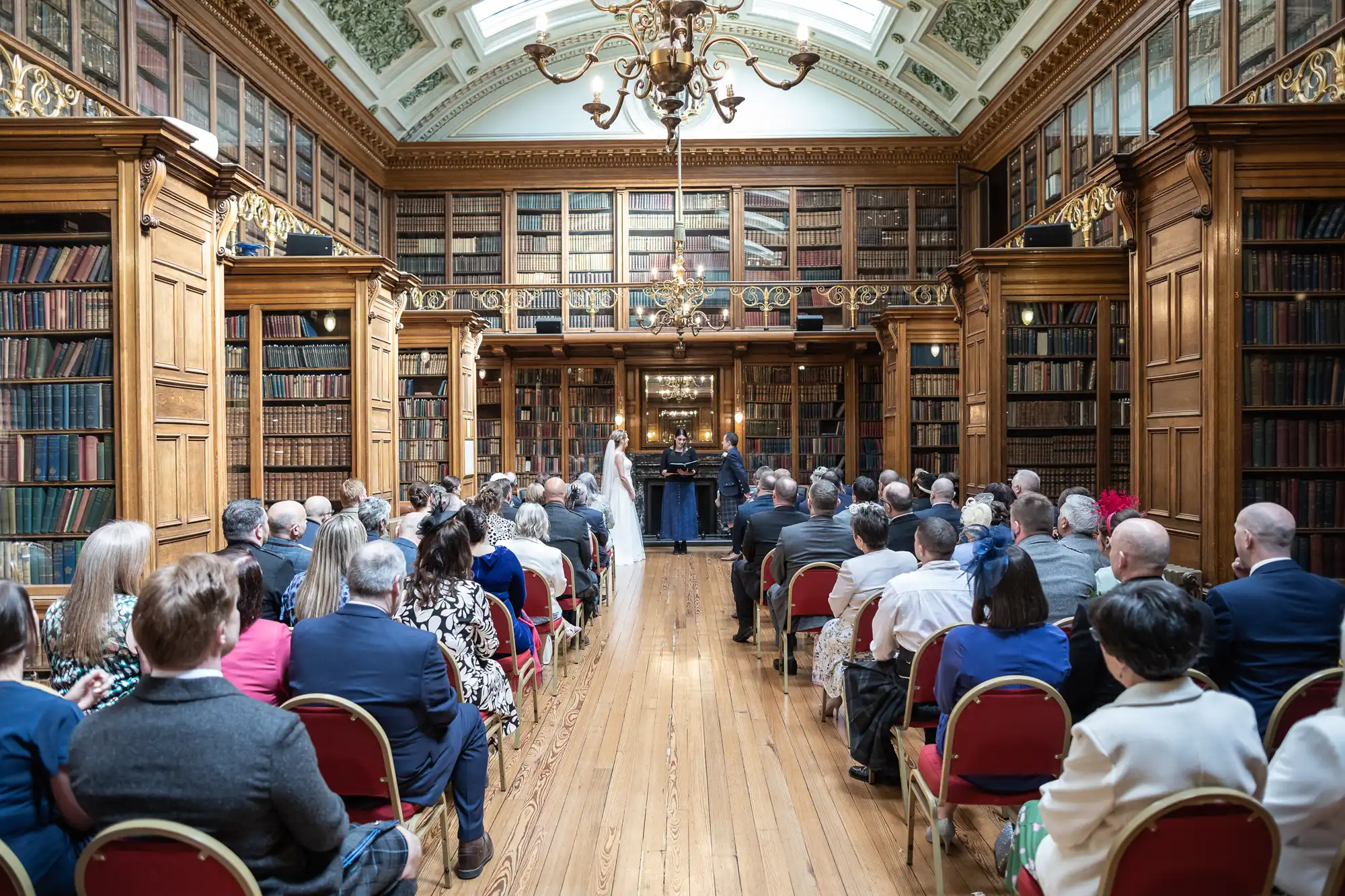 A wedding ceremony taking place in a large, ornate library with rows of seated guests and the bride and groom standing at the front.