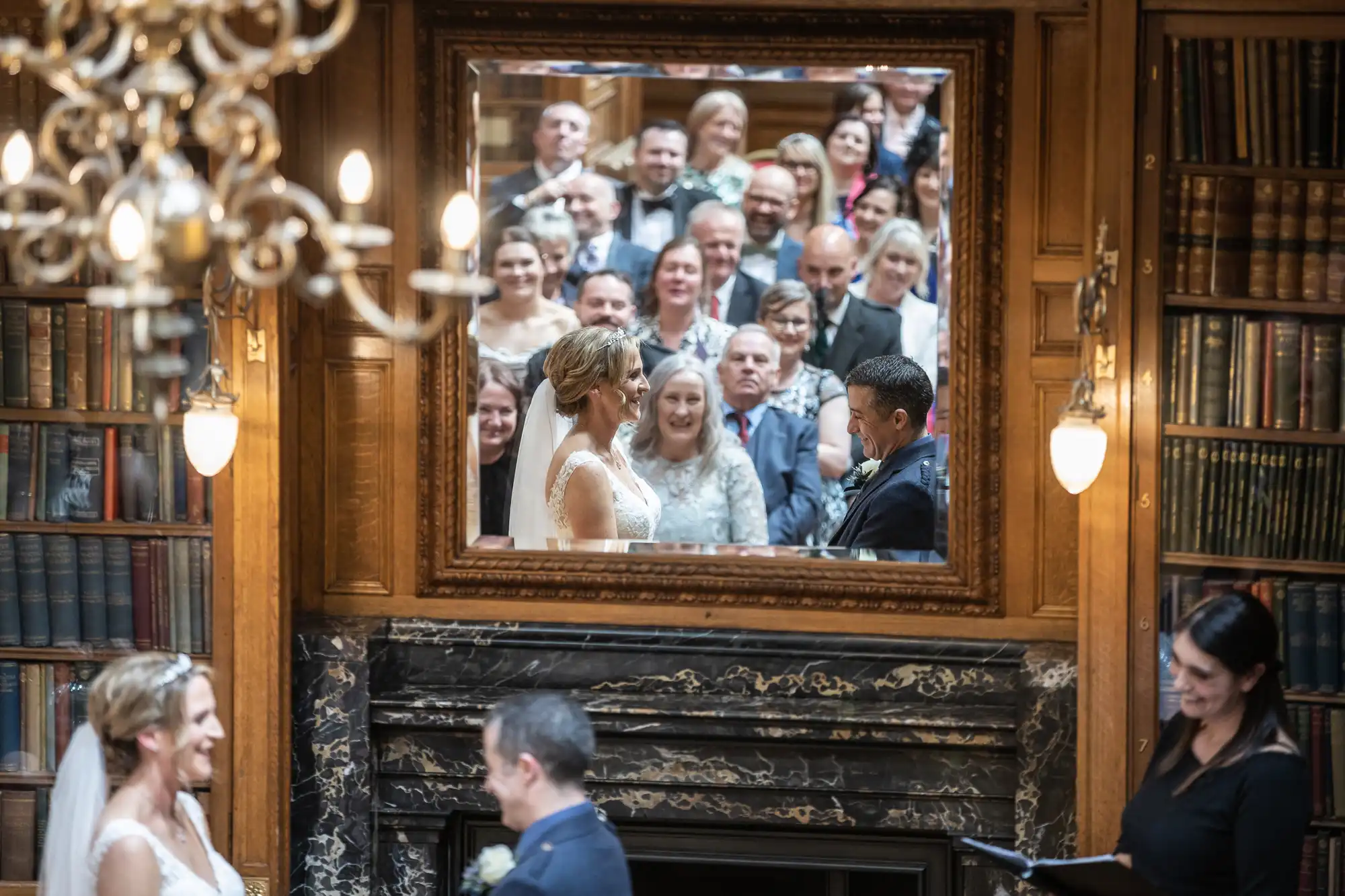 A wedding ceremony with the couple standing at the altar is reflected in a large framed mirror. Guests are seated and watching the ceremony, surrounded by wooden bookshelves and a marble fireplace.