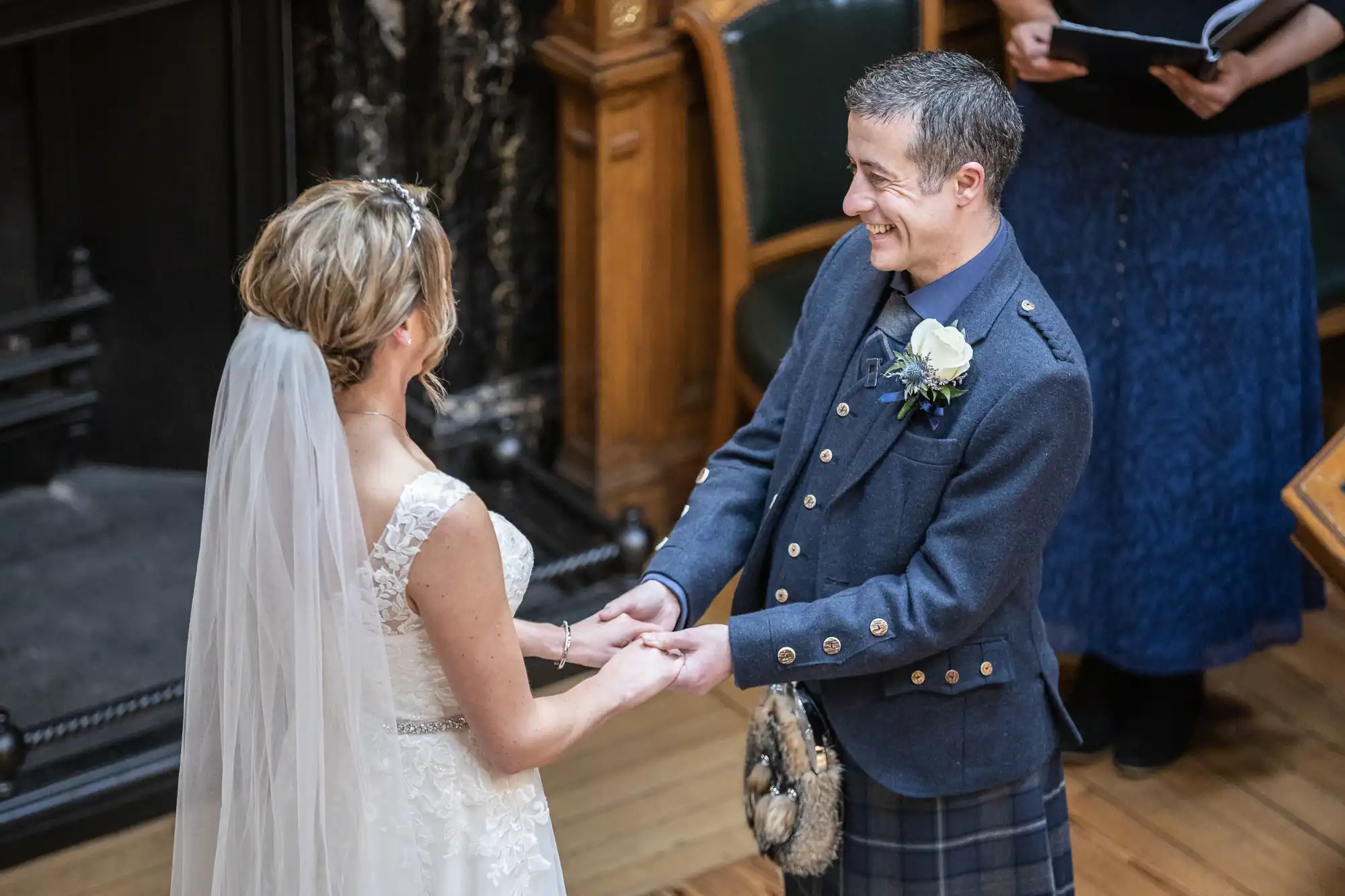 A bride and groom exchange vows and hold hands during their wedding ceremony. The groom wears a traditional Scottish kilt outfit, and the bride is in a white wedding dress with a veil.