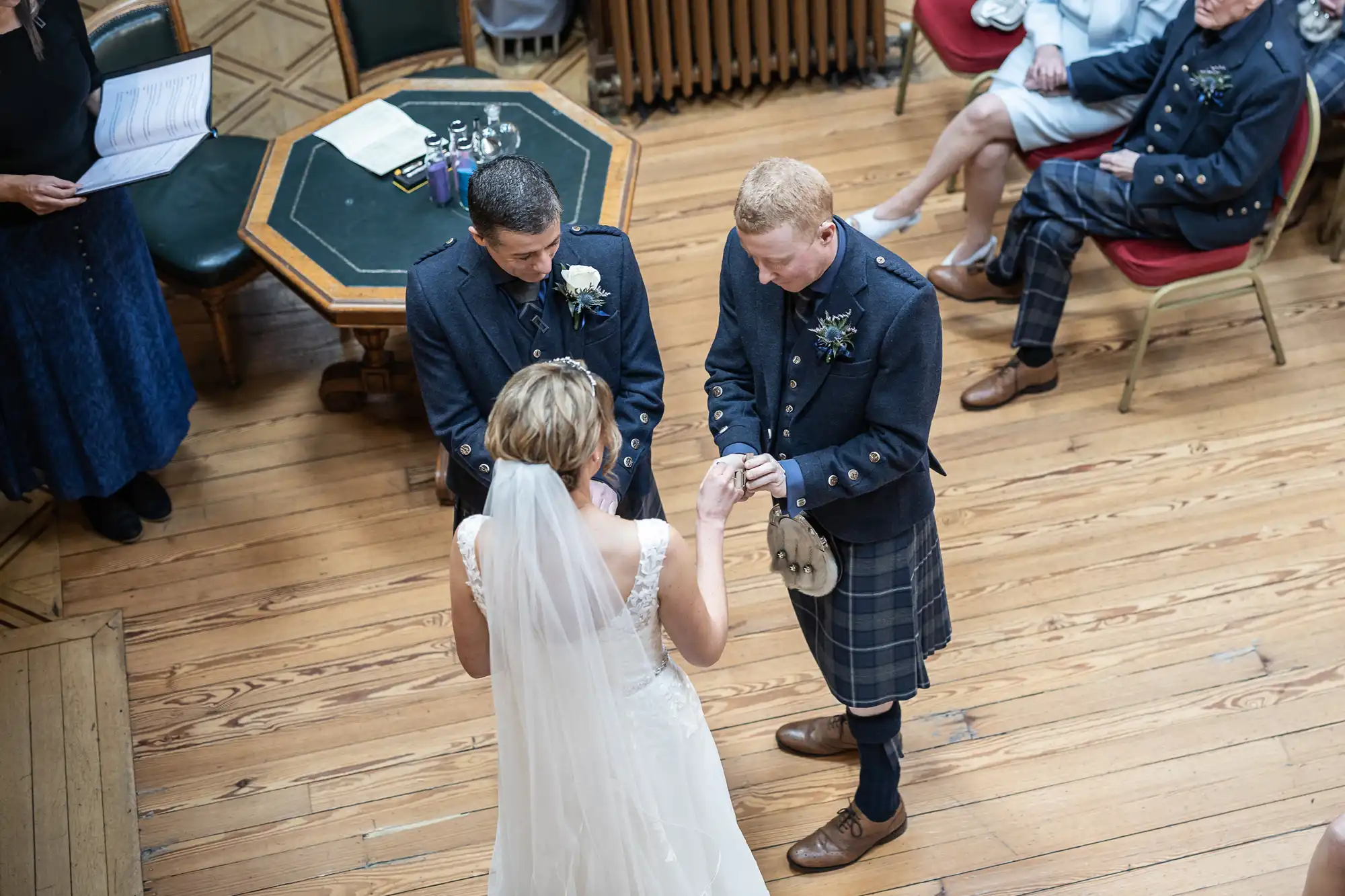 A bride and groom exchange rings during a wedding ceremony. The groom and another man are dressed in kilts, while seated attendees look on.
