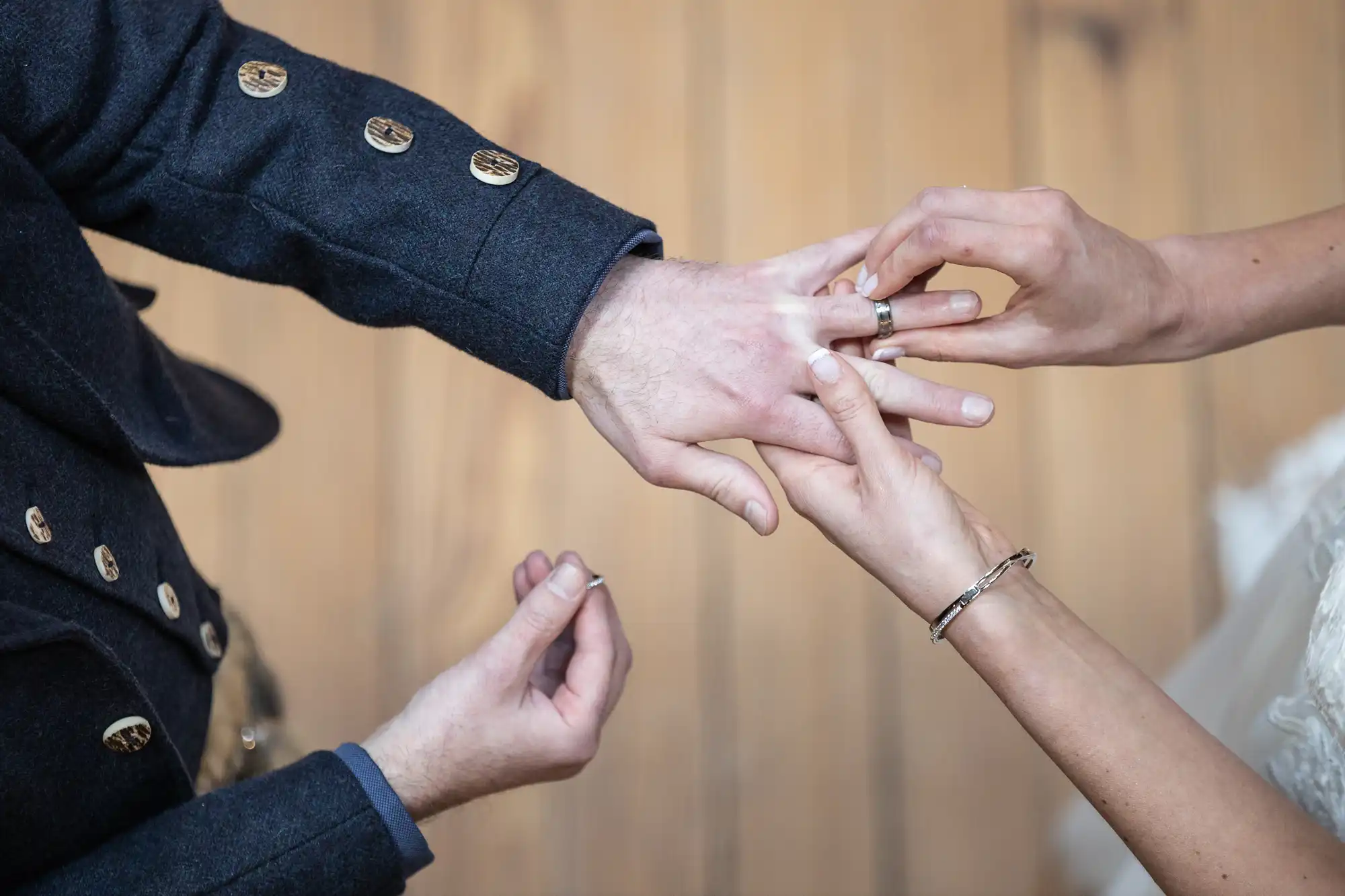 A close-up of a couple exchanging wedding rings. The person on the left is wearing a dark suit, while the person on the right is wearing a bracelet.