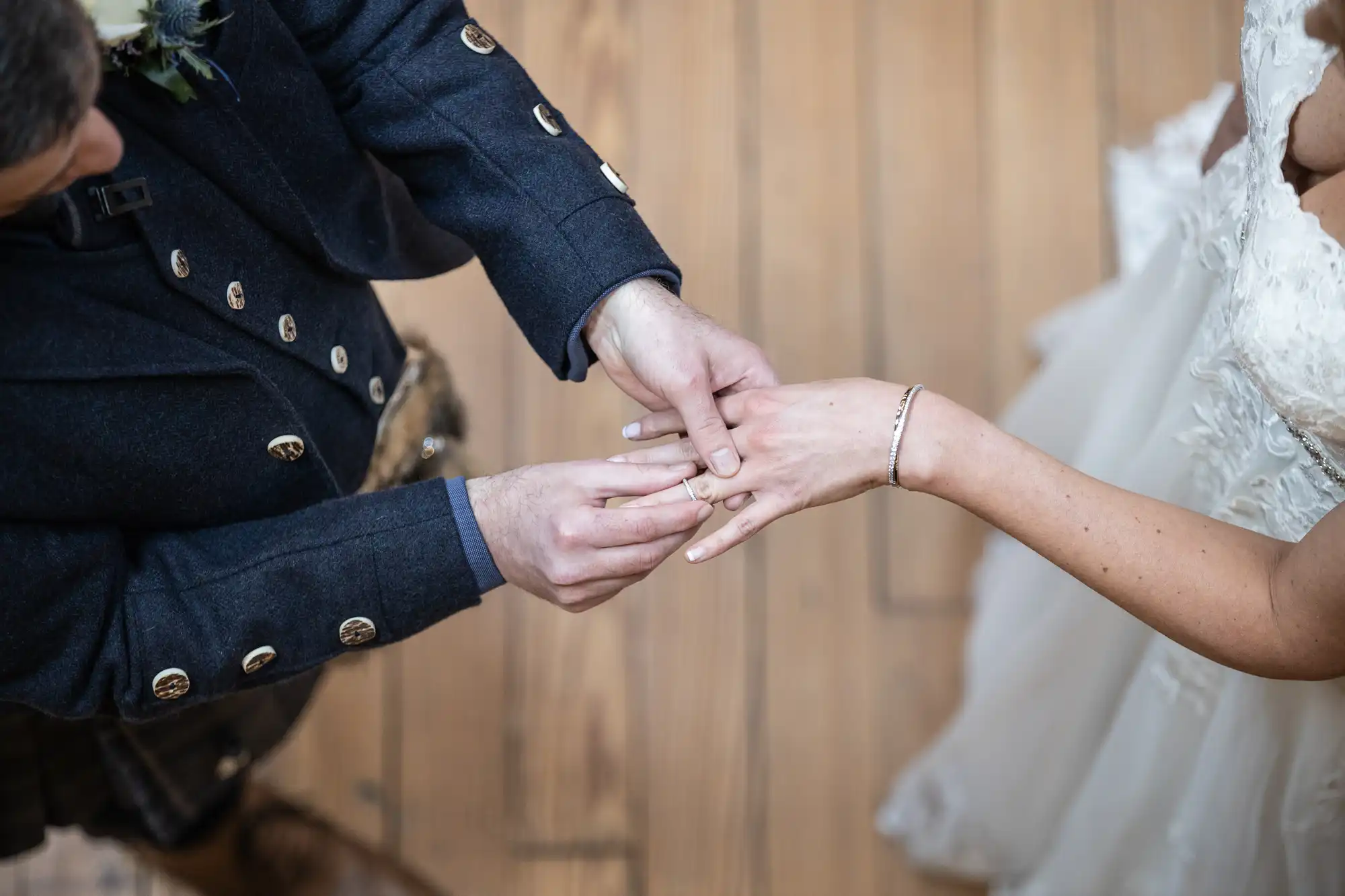 Close-up of a couple exchanging rings during a wedding ceremony. The bride is wearing a white dress, and the groom is in a dark suit with gold buttons. The focus is on their hands and the wedding rings.
