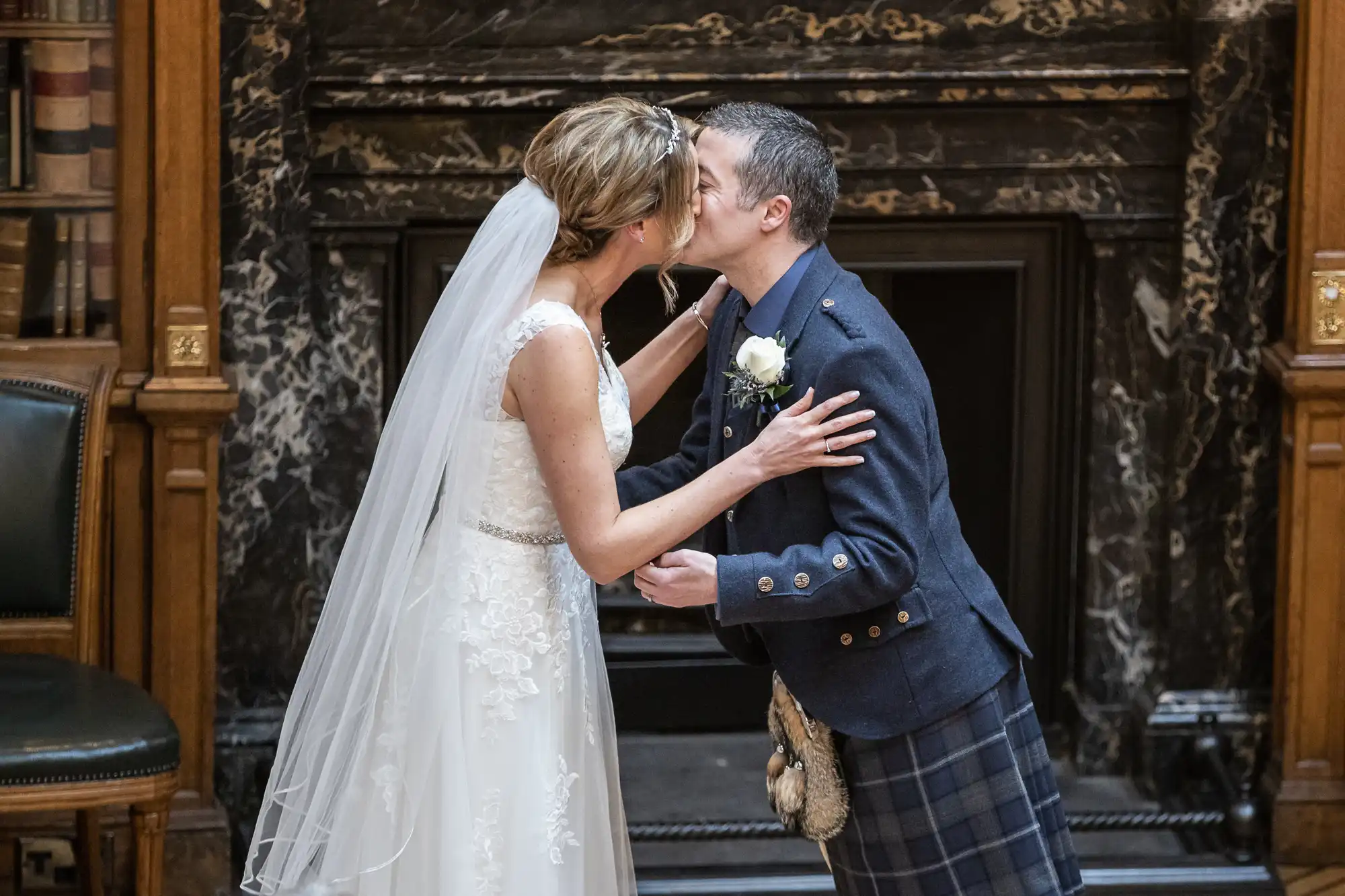 A bride in a white gown and veil kisses a groom in a tartan kilt and dark jacket in front of a marble fireplace.