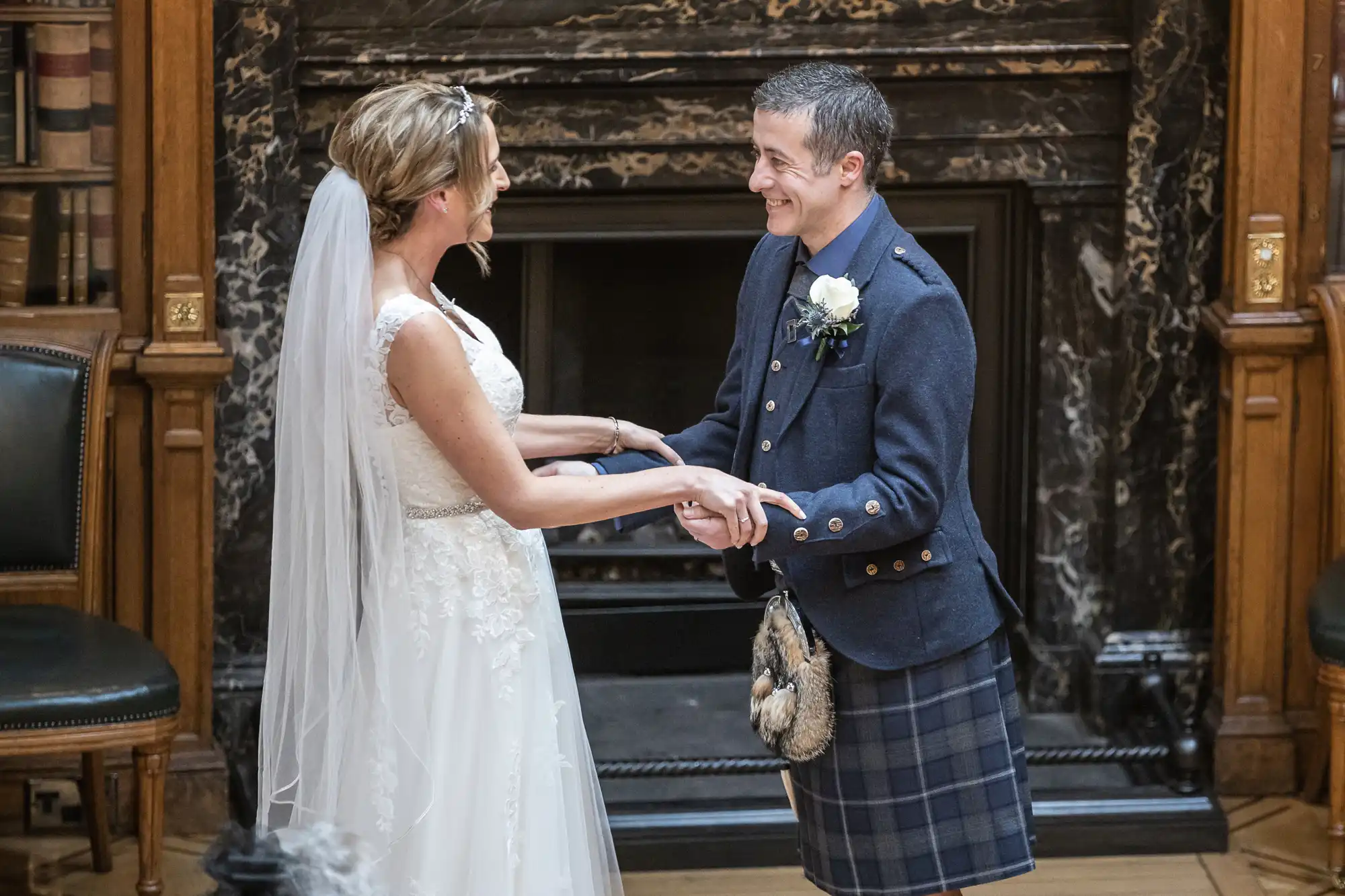 A couple stands, smiling and holding hands, during a wedding ceremony. The bride is in a white dress and veil, and the groom is in a kilt and formal jacket.