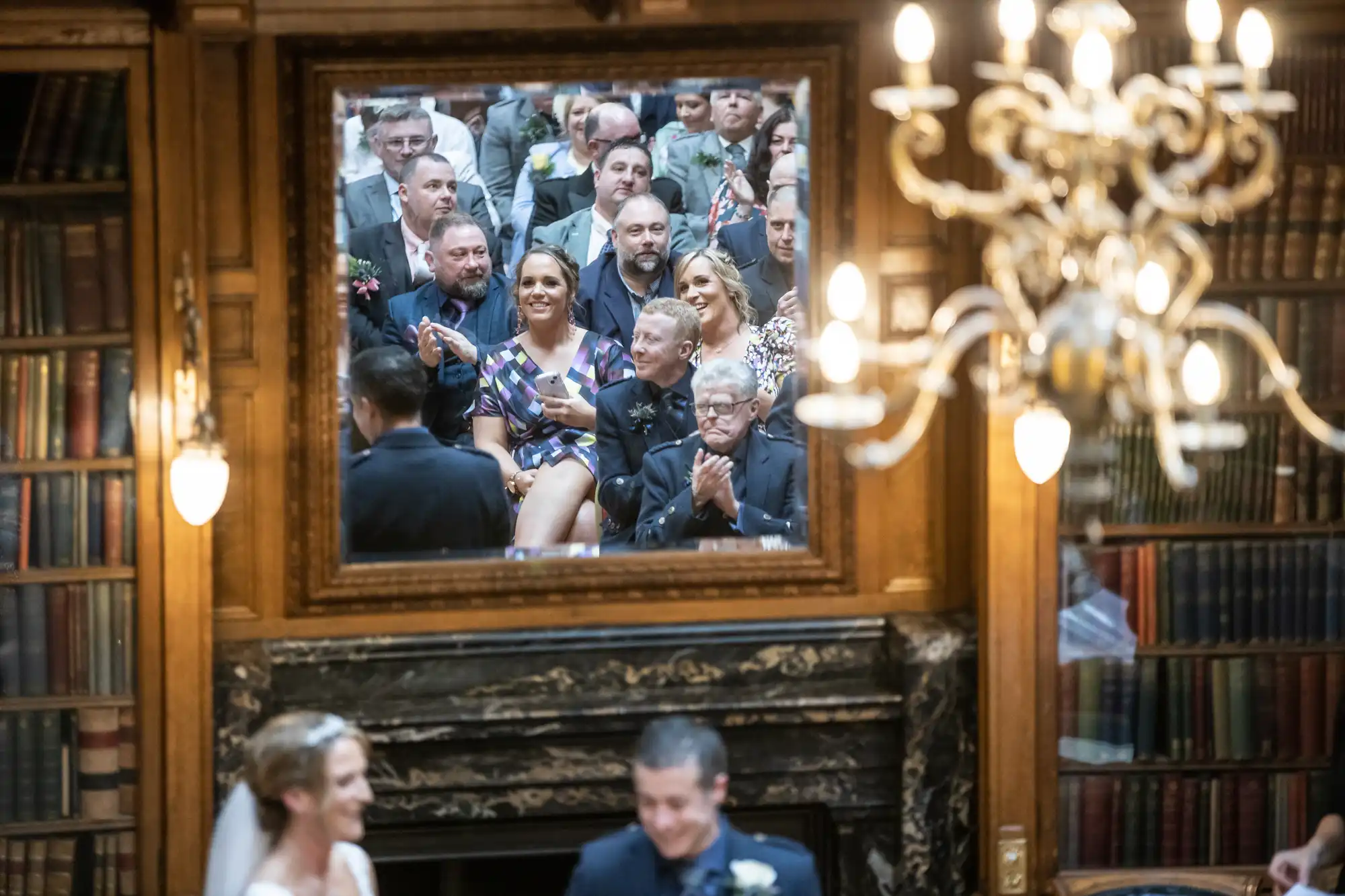 Guests at a wedding are reflected in a large framed mirror in a book-lined, chandelier-lit room, while the bride and groom are blurred in the foreground.
