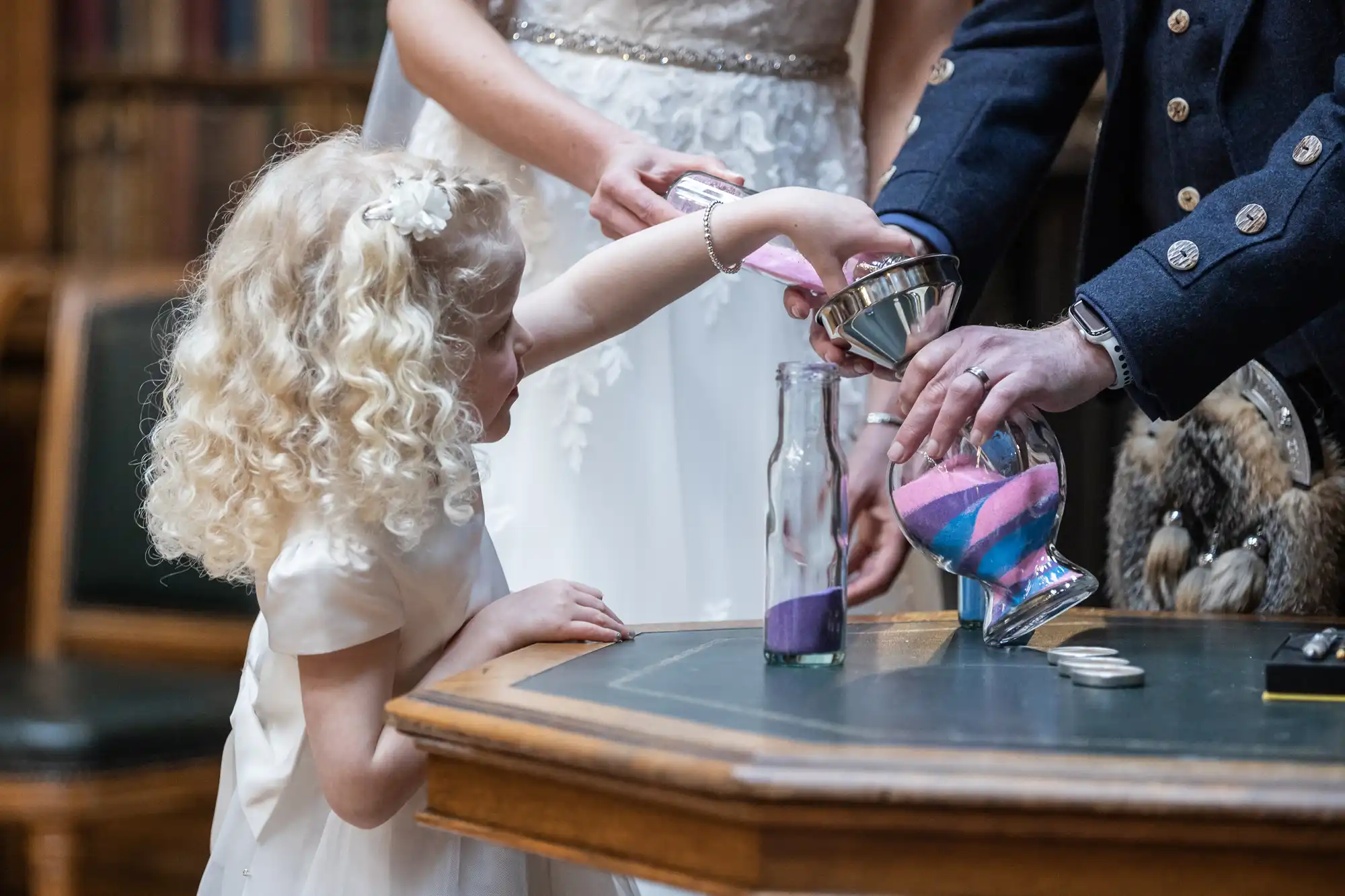 A young girl pours colored sand into a glass bottle, assisted by an adult, while a bride and groom stand nearby.