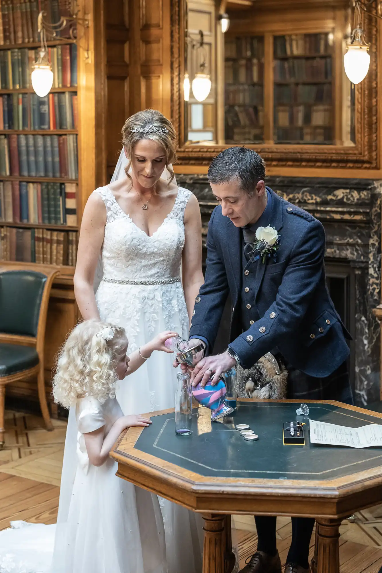 A bride and groom, dressed in wedding attire, are pouring sand into a bottle with a young girl in a formal setting with bookshelves and a fireplace in the background.