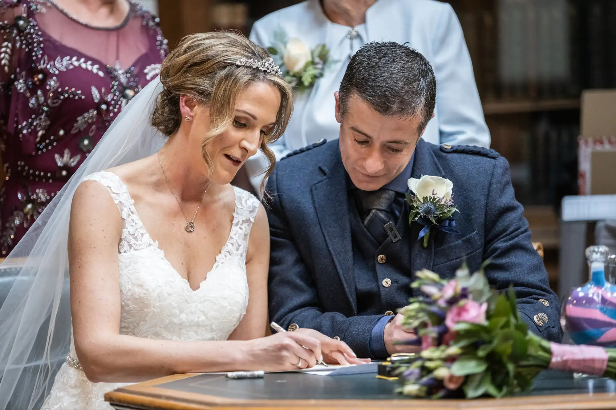 A bride in a white dress and a groom in a dark suit sign documents at a table with a bouquet of flowers.