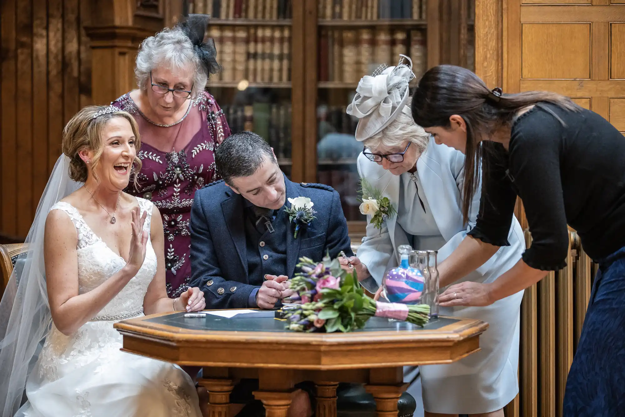 A group of people, including a bride and groom, are seated at a table in a formal setting, possibly a wedding venue. They are signing documents with the help of a woman. Flowers are on the table.