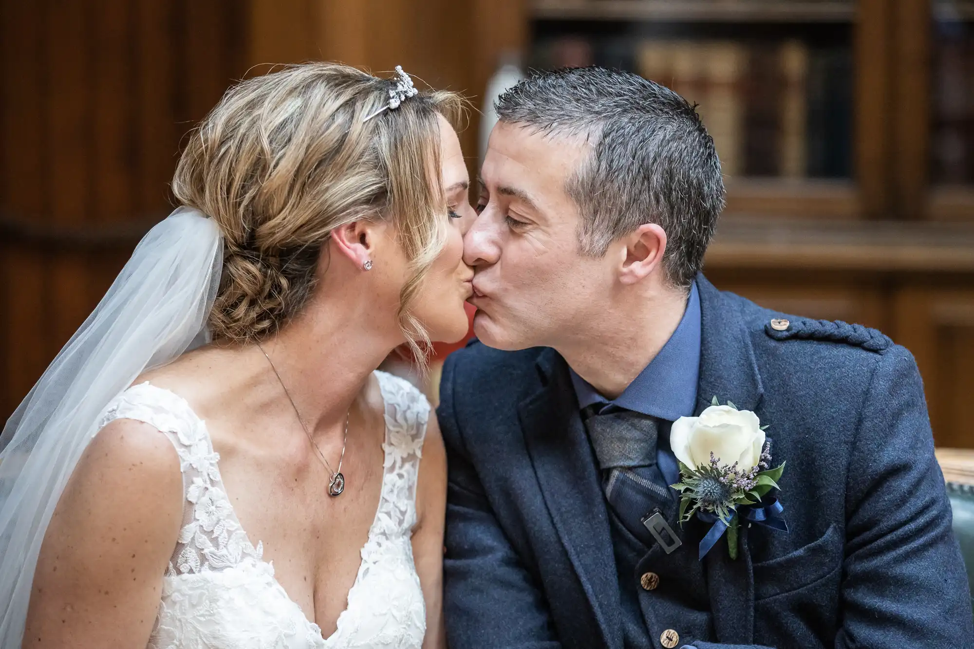 A bride and groom share a kiss. The bride is wearing a lace wedding dress and veil, and the groom is wearing a suit with a boutonniere. Both have closed eyes and look happy.