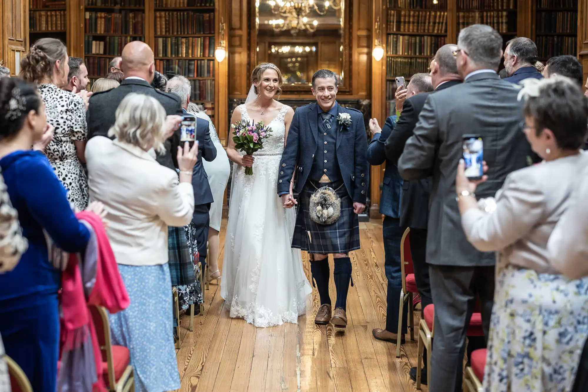 A newly-married couple, with the groom in a kilt and the bride in a white dress, walk down the aisle as guests take photos in a library setting.