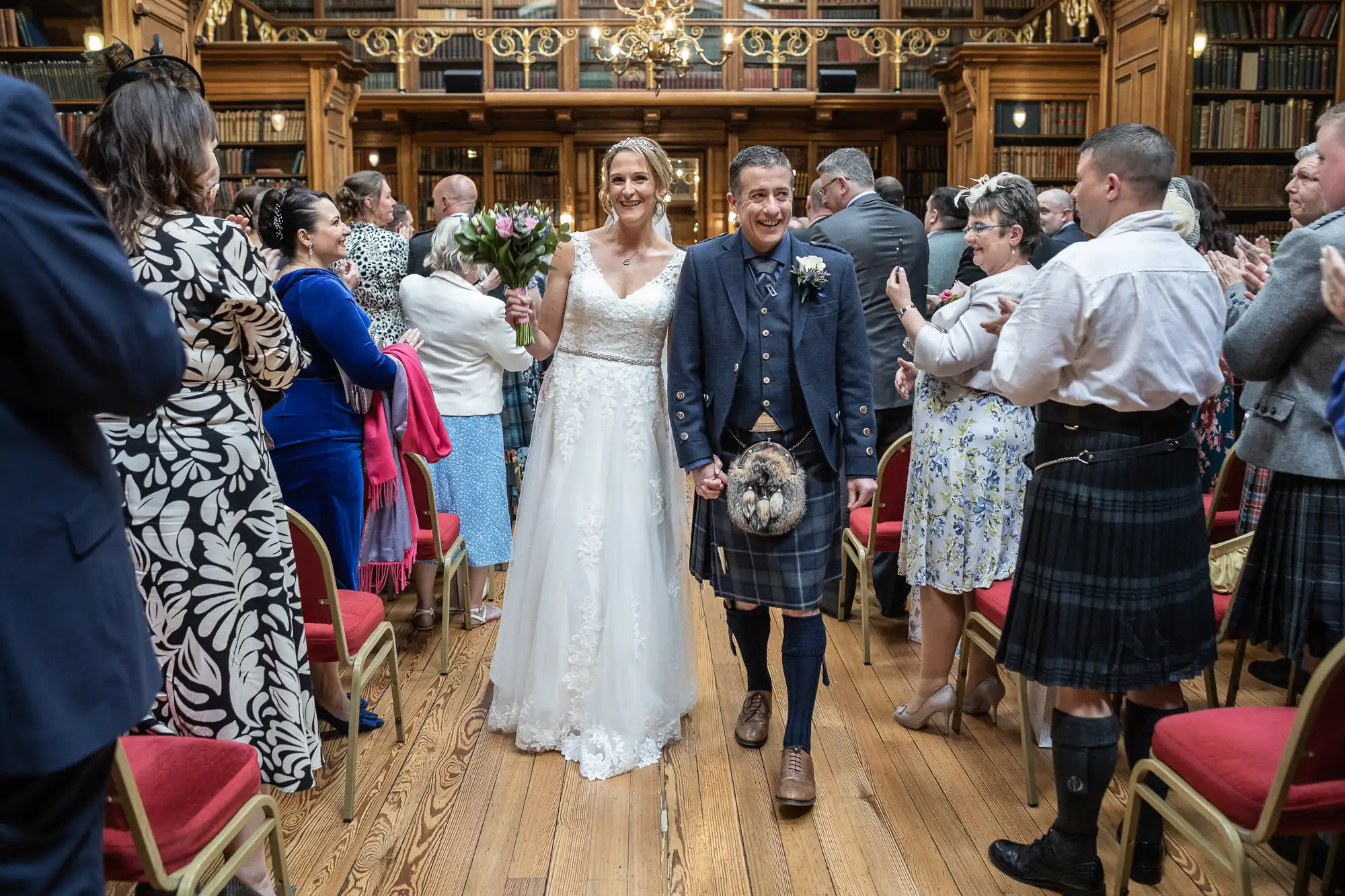 A bride in a white dress and groom in a kilt walk down the aisle, surrounded by applauding guests in a wood-paneled room with bookshelves and ornate decor.