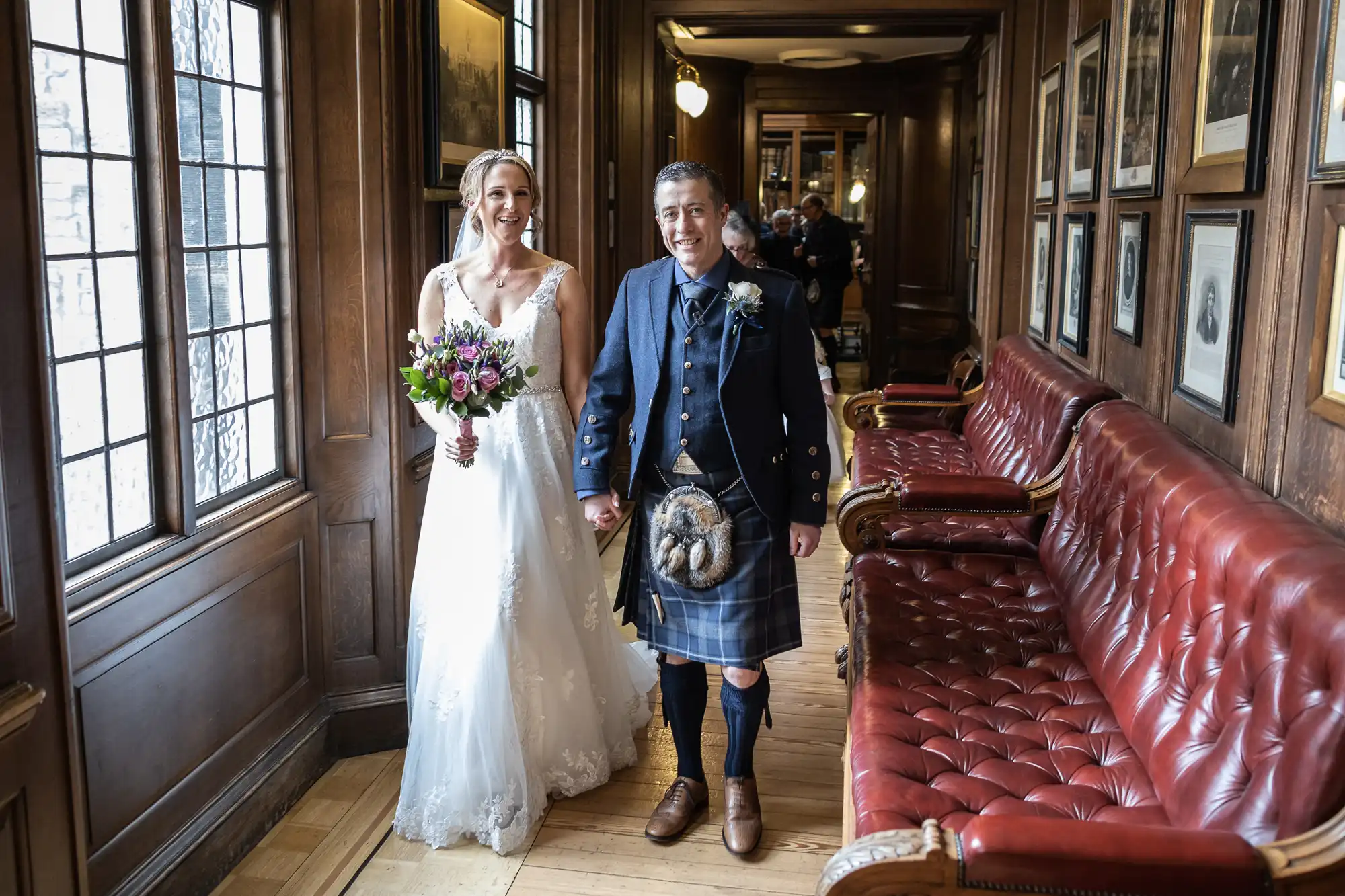 A bride in a white gown and a groom in traditional Scottish attire, including a kilt, walk hand in hand down a wood-paneled hallway lined with framed pictures and red leather benches.