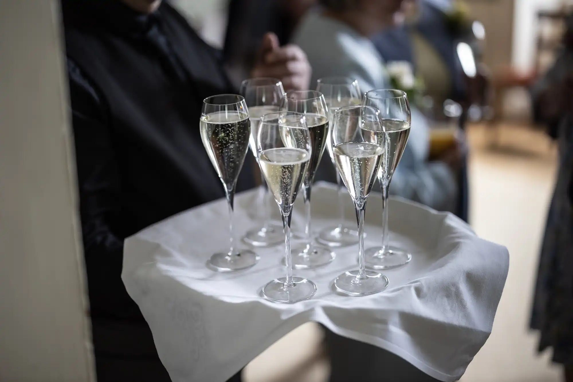 A person holding a tray with eight filled champagne flutes in a social event setting.
