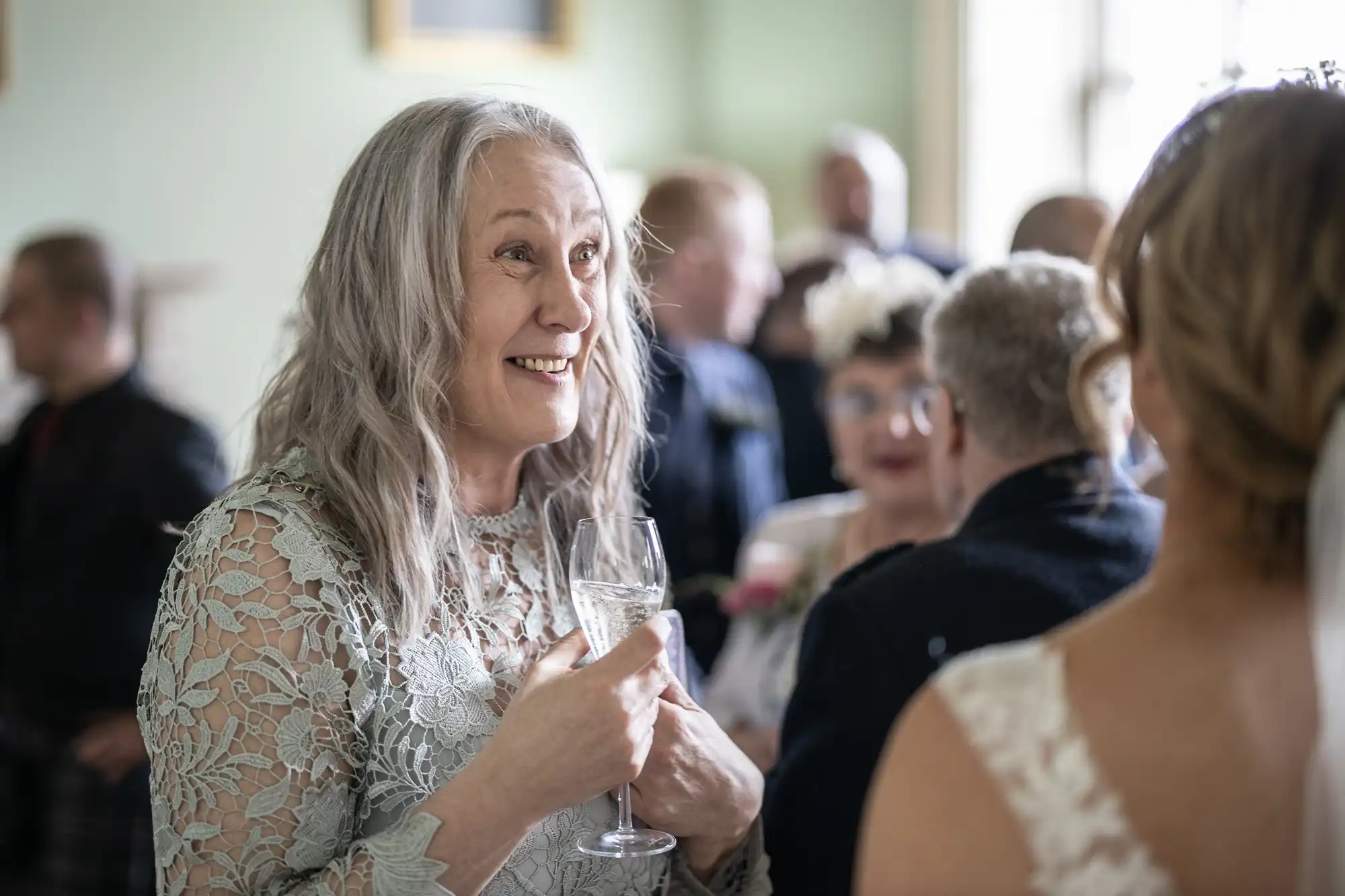 An elderly woman with long gray hair, wearing a lace dress, holds a glass of wine and smiles while talking to another person at an indoor gathering.