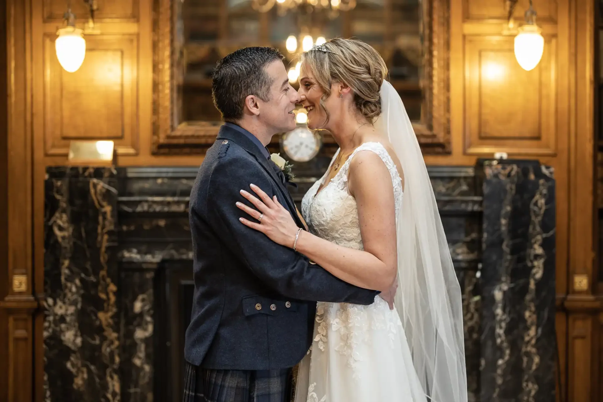 A bride and groom smile at each other, standing in front of a marble fireplace with wooden paneling and lights on either side. The bride wears a white dress and veil, and the groom is in a dark suit.