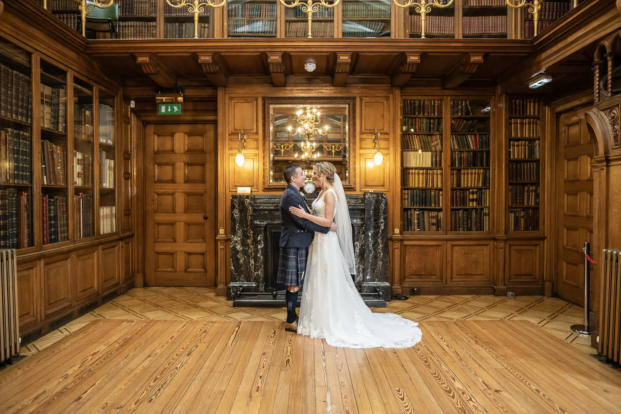 A bride and groom, dressed in a white wedding gown and a kilt, share a kiss in a grand wood-paneled library filled with bookshelves.