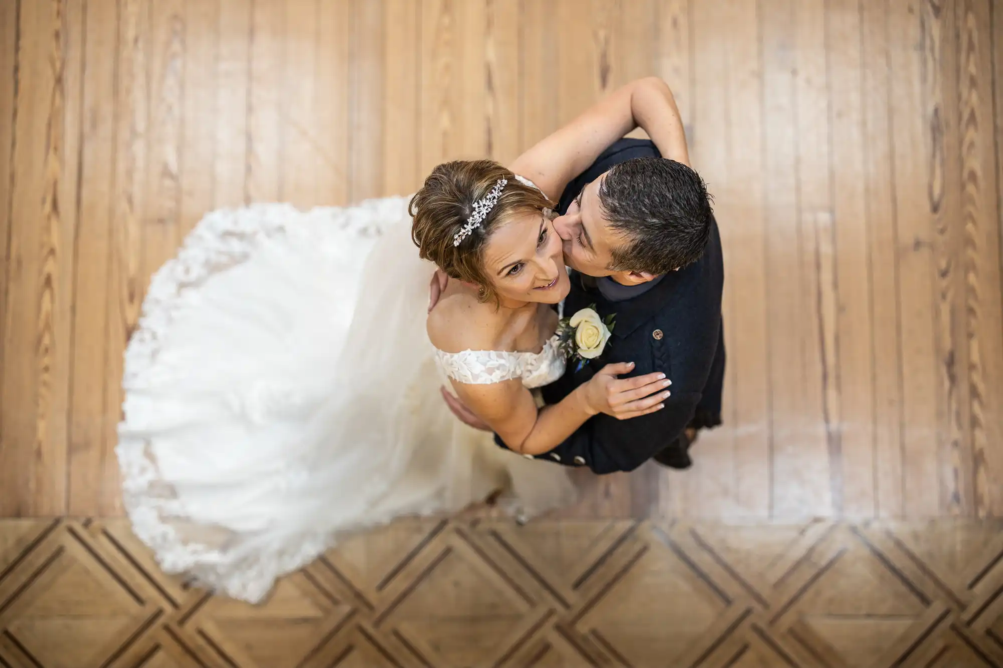A bride and groom share a dance on a wooden floor, captured from above. The bride wears a white dress and headpiece, while the groom is in a dark suit with a boutonnière.