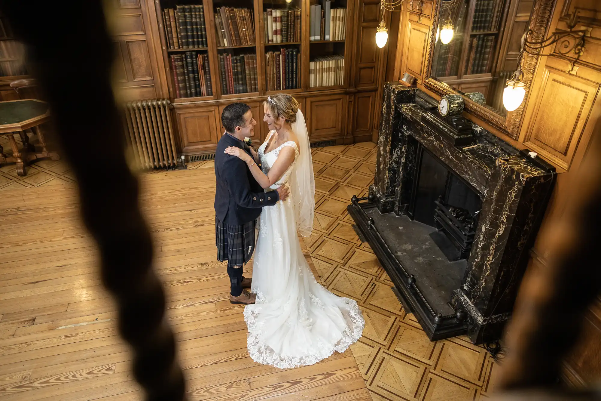 A bride and groom share a dance in a wood-paneled library, lit by ornate hanging lights. The groom wears a kilt, and the bride is in a white gown and veil.