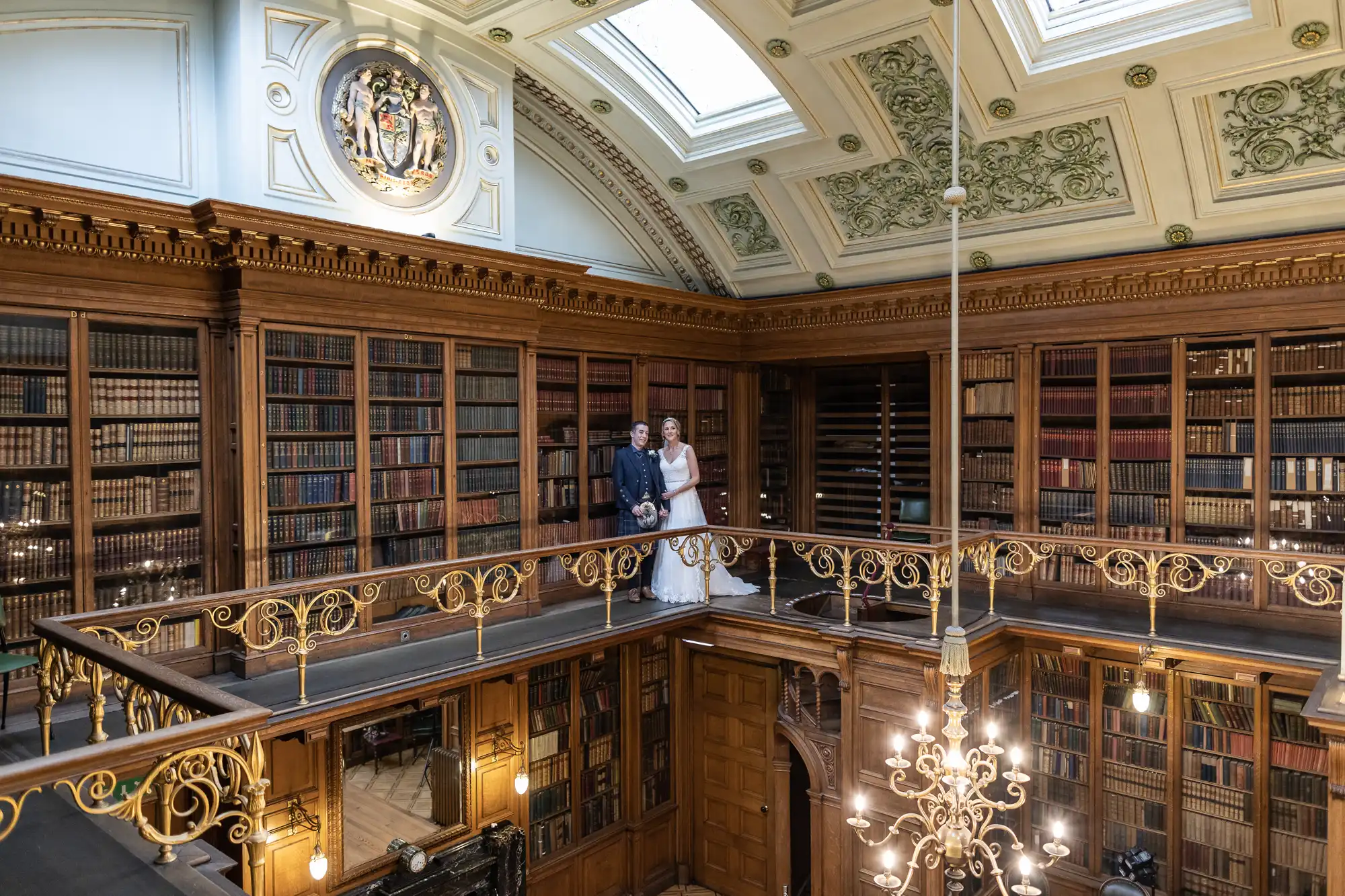 A couple stands on a balcony inside a grand, wood-paneled library with tall bookshelves and ornate ceiling details.