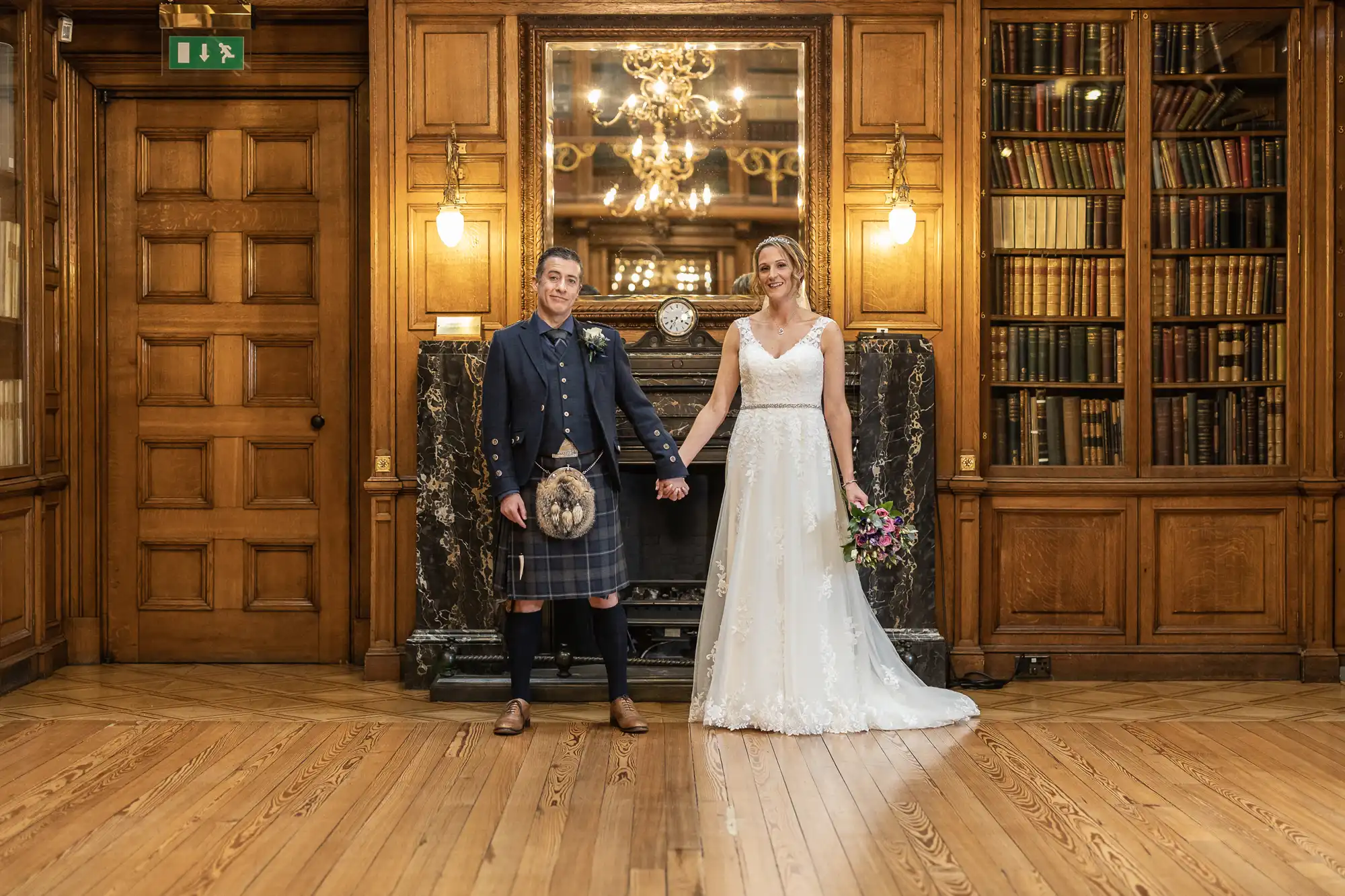 A couple in wedding attire stands in a wood-paneled room filled with bookshelves and a fireplace. The man wears traditional Scottish dress, and the woman is in a white gown holding a bouquet.