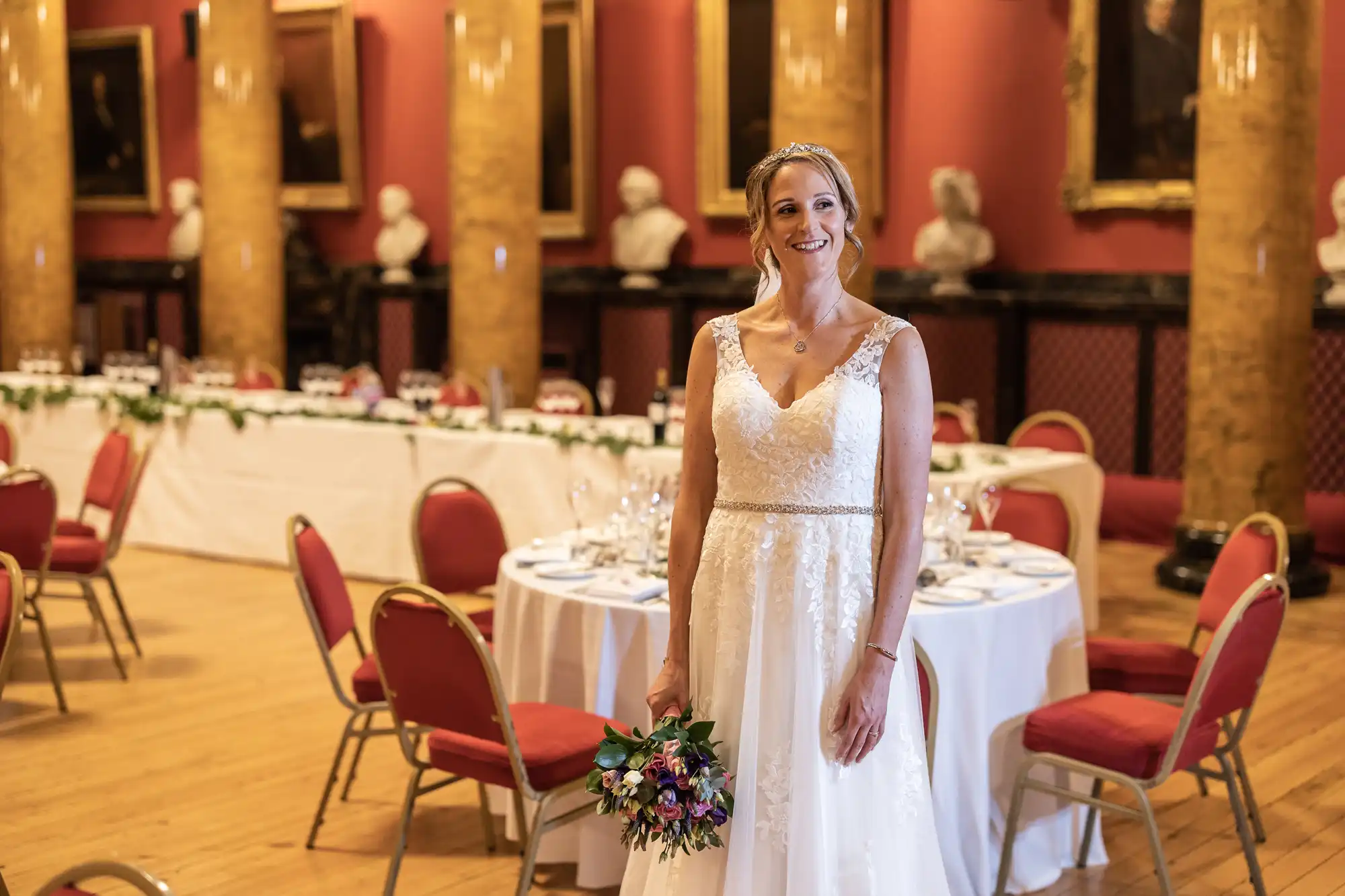 A bride in a white wedding dress stands smiling, holding a bouquet in a hall with red chairs and a long table decorated for a reception.