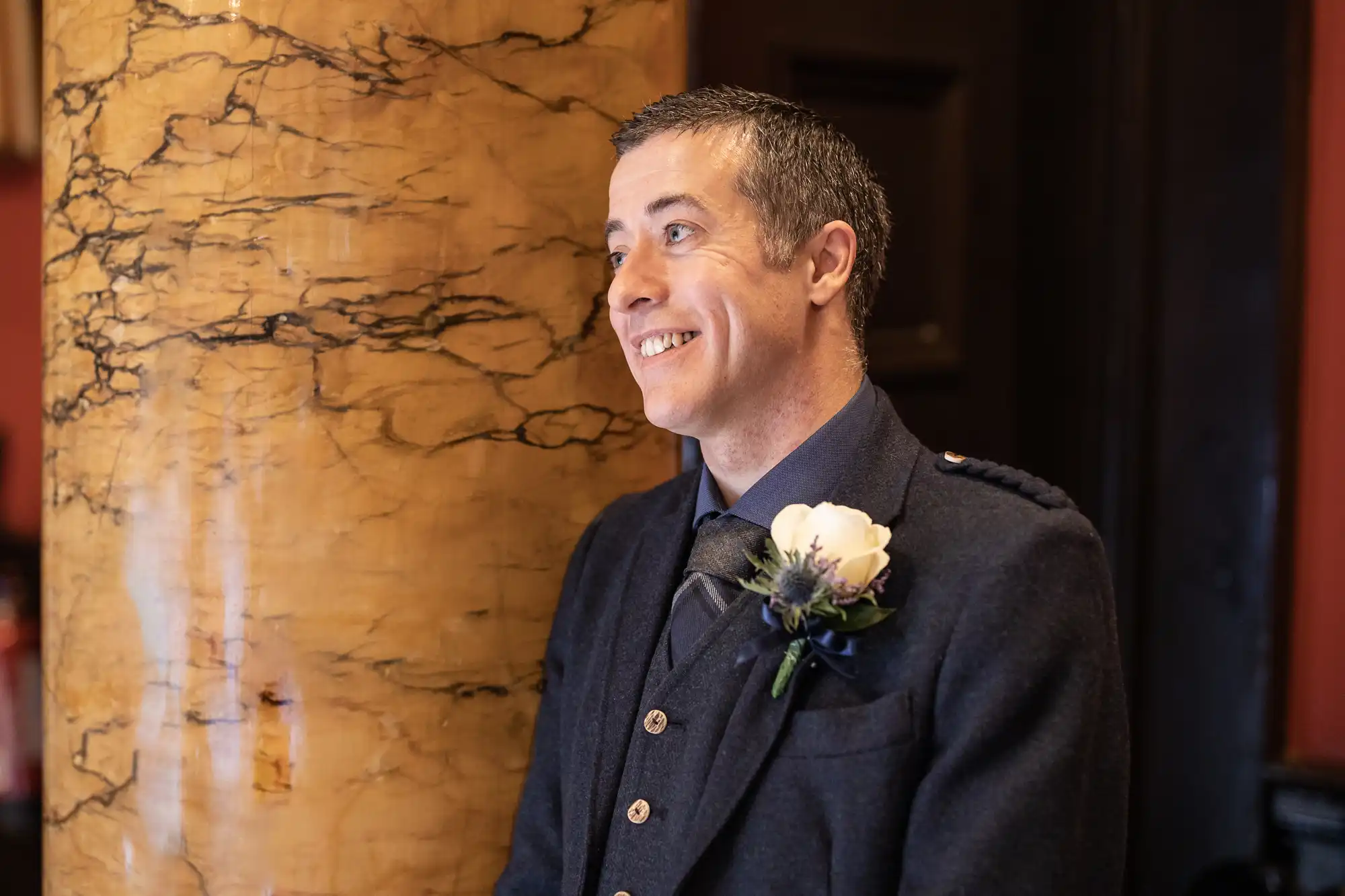 A man in formal attire with a boutonniere smiles while standing next to a marble column.