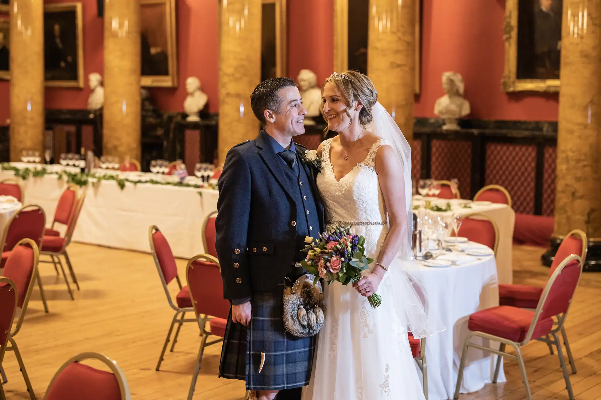 A couple, dressed in wedding attire, stand smiling at each other in a grand room with red walls, statues, and set dining tables. The groom wears a kilt and the bride holds a bouquet of flowers.