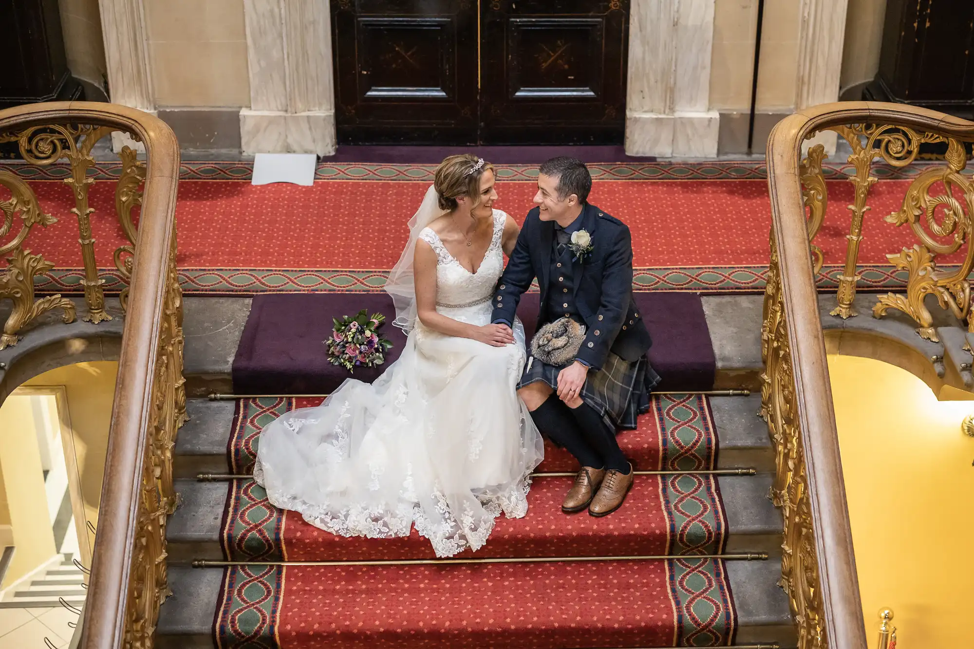 Bride and groom sitting on ornate stairs. Bride wears a white gown, groom wears a kilt. Bouquets of flowers are placed beside them. Jenna is leaning into Tom in this moment of shared happiness.